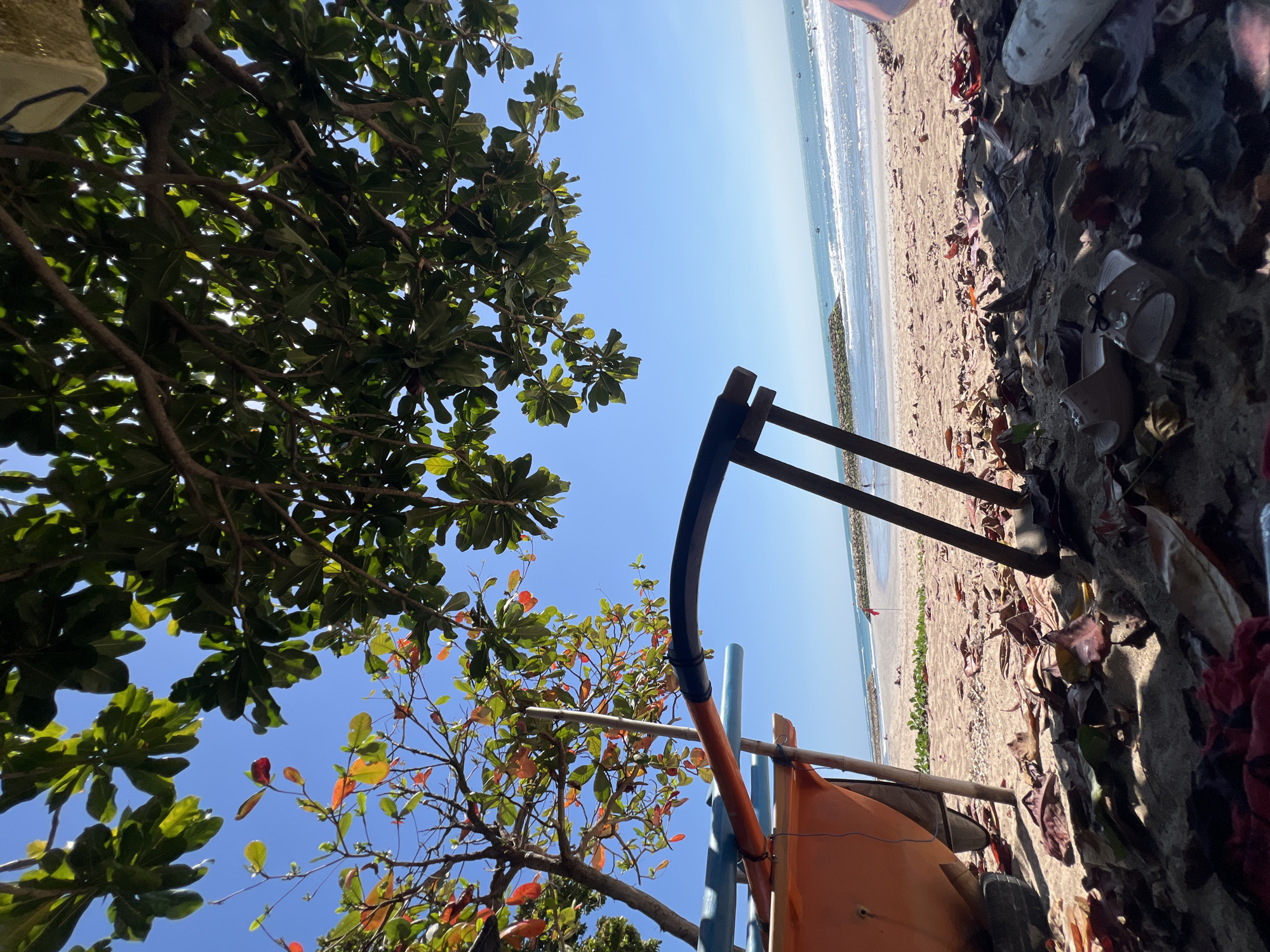 The image depicts a low-angle view of a beach in Kuta, Indonesia, on a clear, sunny day. The foreground is dominated by fallen leaves scattered on the sandy ground, with a pair of sandals visible among them. A partially visible orange boat, supported by wooden poles, is positioned to the left. Two dark, curved metal poles extend upwards from the sand, possibly part of a beach umbrella or seating structure. In the mid-ground, the sandy beach meets the gentle waves of the ocean. A breakwater, constructed of rocks, extends into the sea, and a small red flag is planted on the beach. The background features a clear blue sky with lush green foliage from overhead trees obscuring parts of the sky and casting shadows. The overall scene suggests a quiet moment on the beach, possibly in the late morning or early afternoon.