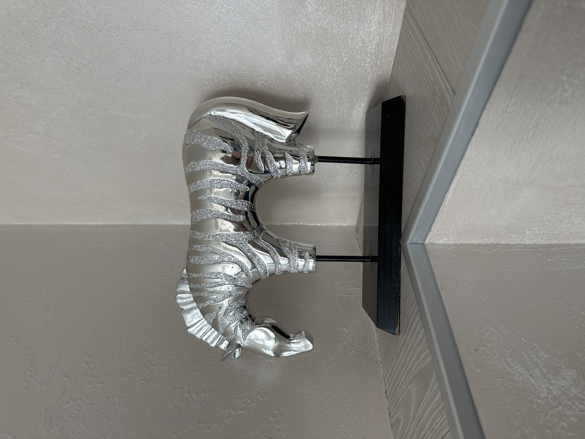 An art deco-style silver zebra sculpture with glittery stripes stands on a black rectangular base, supported by two thin black rods. The sculpture is positioned on a light-colored wooden shelf in a corner where two beige walls meet. A reflective gray trim runs along the front edge of the shelf. The background features textured beige walls.