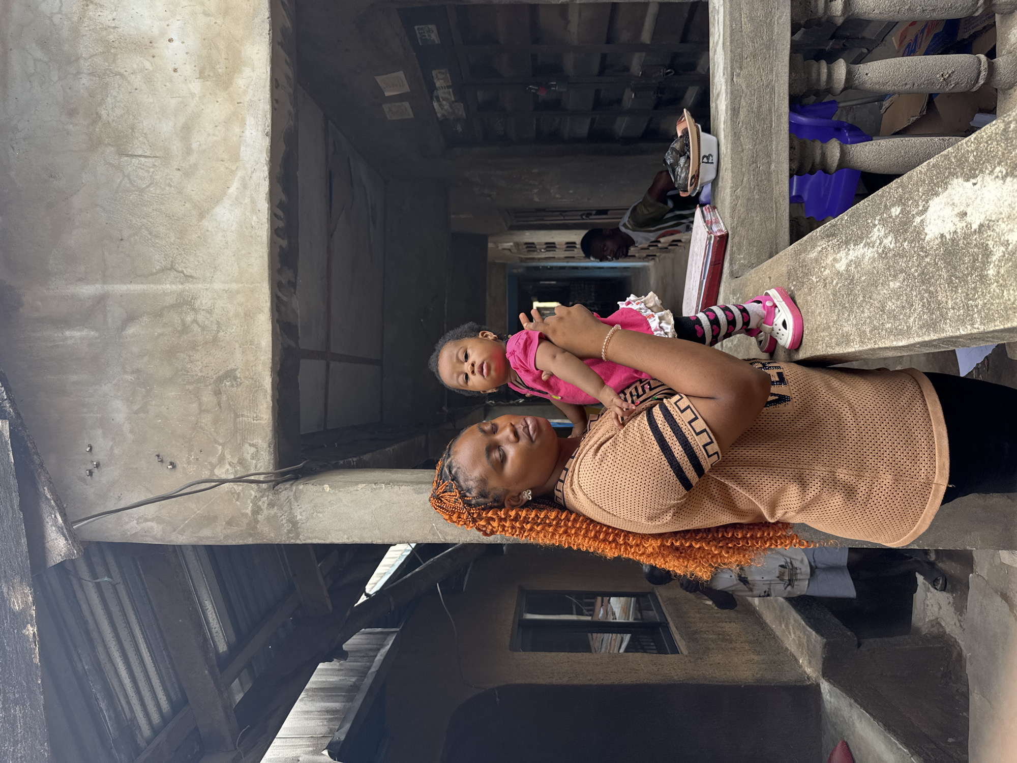 A woman stands on a concrete balcony in Onitsha, Nigeria, holding a young child. The woman has orange braided hair and wears a tan, perforated top with black accents, black pants, and small earrings. The child wears a pink top, striped leggings, and pink and white shoes. Behind them, a dark corridor reveals a glimpse of another person. The background includes deteriorating concrete structures, exposed wiring, and corrugated roofing, typical of an urban environment. Concrete balustrades, boxes, and a white hat are visible on the balcony.