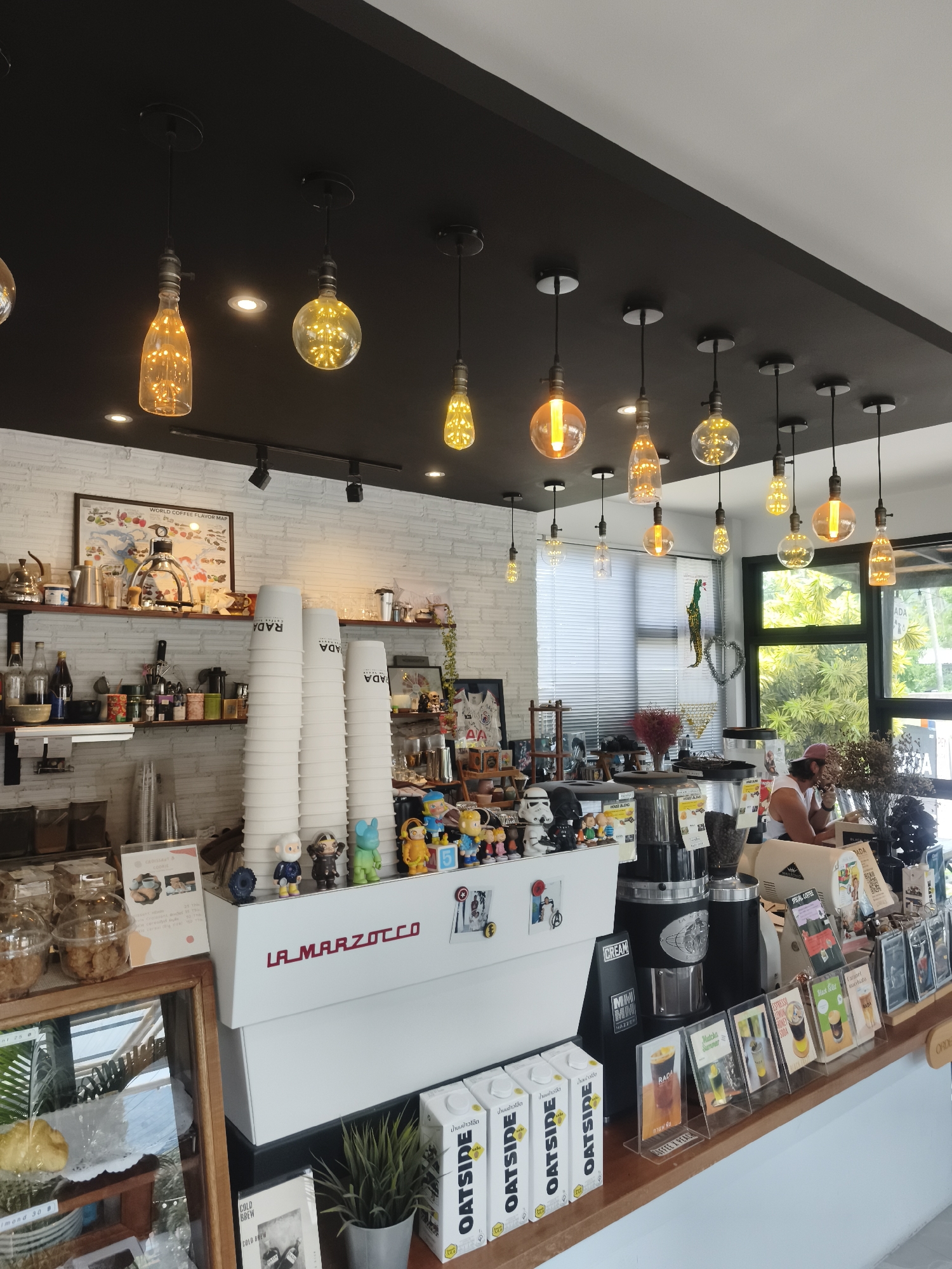 The image depicts the interior of a modern coffee shop in Ko Samui District, Thailand. A white La Marzocco espresso machine is prominently displayed on the counter, with stacks of white paper cups to its left. Various small figurines, including what appear to be cartoon characters and stormtrooper figures, are arranged on top of the machine.
Behind the counter, shelves are stocked with glassware, bottles, and containers. A framed "World Coffee Flavor Map" hangs on the white brick wall. The ceiling features a black panel with numerous exposed filament light bulbs in various shapes, creating a warm ambiance.
In the background, a person wearing a white tank top and a pink baseball cap is seated at the counter, appearing to interact with a drink or a phone. Large windows reveal lush green foliage and trees outside, suggesting a tropical setting. The overall scene conveys a clean, contemporary, and inviting atmosphere.