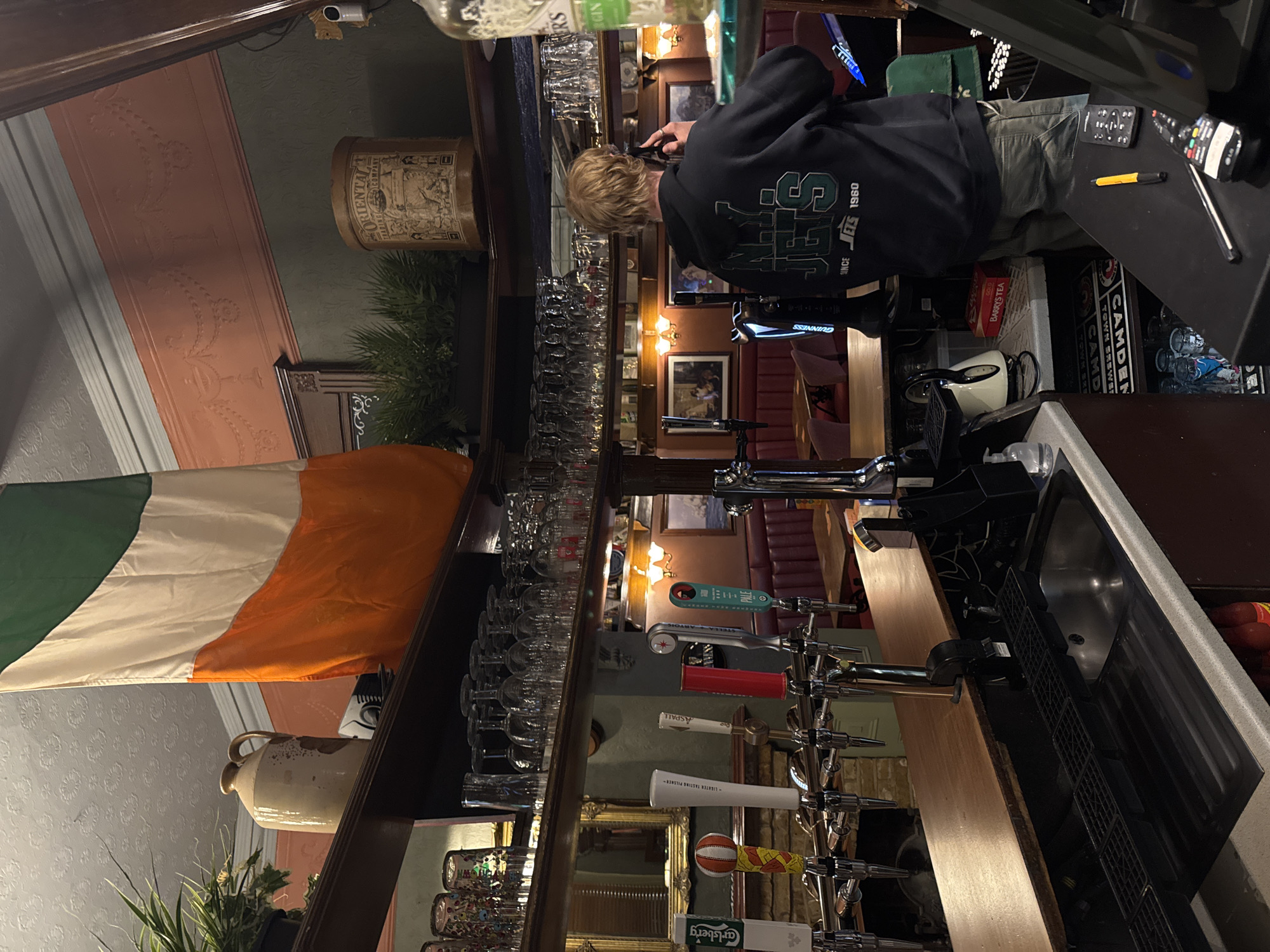 The image captures the interior of a traditional pub or bar, likely in London, United Kingdom, given the visible cues. The scene is dominated by a long, well-stocked bar counter and a person standing behind it.
The person, seen from the back, has short, light brown hair and is wearing a dark blue or black hoodie with a green and white logo that reads "JETS SINCE JETS 1960". They appear to be engaged in an activity behind the bar, with one hand seemingly near their ear, though the exact action is unclear. They are wearing light-colored trousers.
The bar features numerous beer taps along its length, including identifiable handles for "GUINNESS", "STELLA ARTOIS", "ASPALL GOLD", and one marked "PALE". Another tap handle is themed with a basketball pattern, and two others display "LIGHTER TASTING PILSNER" and "Carlsberg". Rows of clean pint glasses and wine glasses are neatly arranged on shelves above and below the bar. On the counter, various items are visible, such as a bar sink, two remote controls, a yellow pen, a kettle, and a box of "BARRYS TEA". Partially visible behind the person is a crate or box with "CAMDEN TOWN BREWERY" text.
Decorative elements contribute to the pub atmosphere. An "ORIENTAL TRADING COMPANY" cylindrical tin rests on a shelf, along with a plant. An Irish flag, with its green, white, and orange stripes, hangs prominently on the left side of the frame. The walls behind the bar have patterned wallpaper in tones of green and pink, with upholstered red seating visible further back. Artificial lighting from wall sconces and overhead fixtures illuminates the space, suggesting it is evening or indoors.