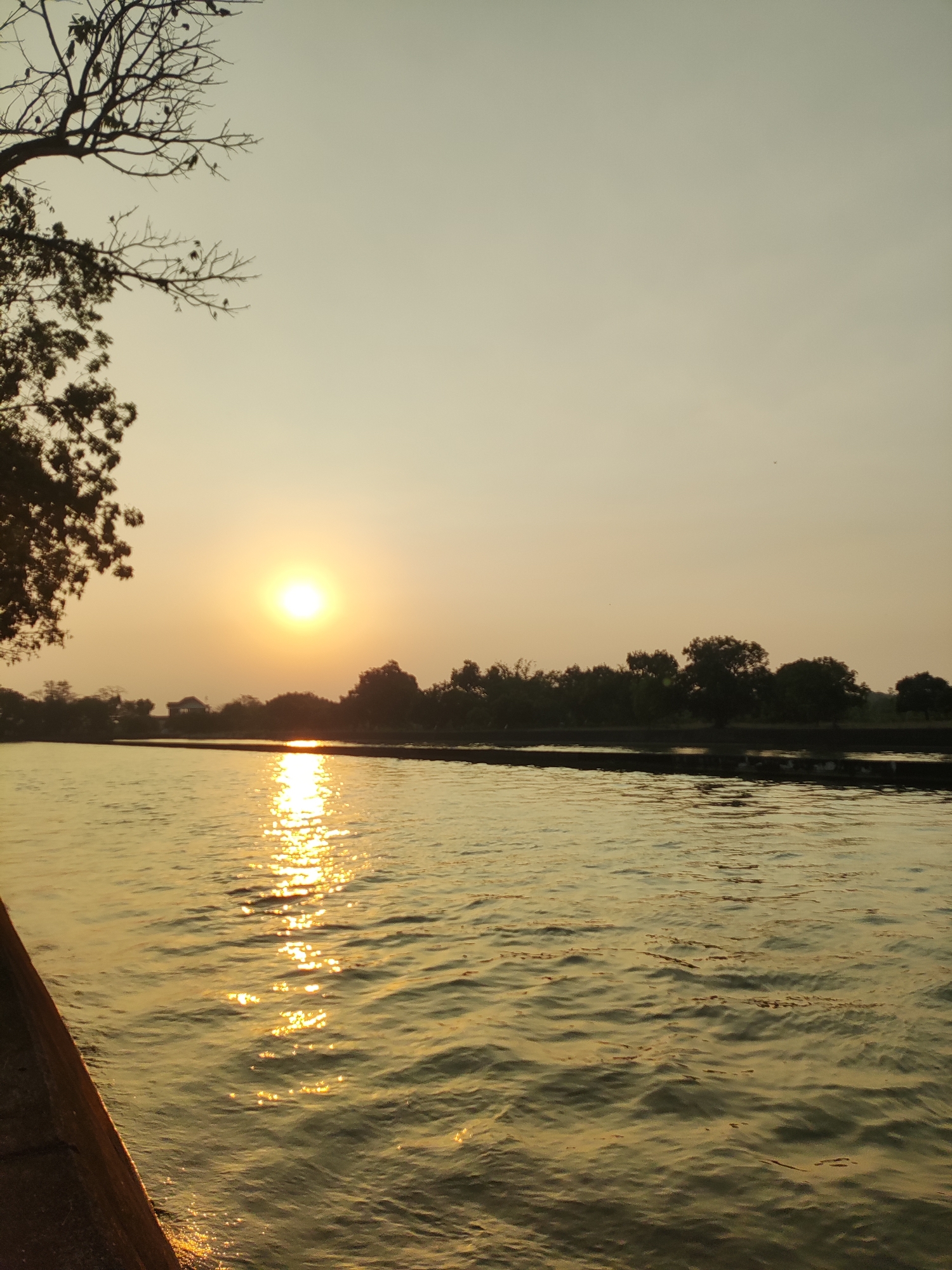 The image depicts a tranquil sunset scene over a body of water in Beber, Indonesia. The sun, a bright orange orb, is low on the horizon, casting a warm, golden glow across the sky and reflecting shimmeringly on the gently rippling water. Silhouetted trees and a small building form the distant landscape along the far bank of the water. In the foreground, the edge of a concrete embankment is visible on the left side of the frame, leading the viewer's eye into the scene. The water appears calm, with subtle waves catching the sunlight. The overall atmosphere is serene and peaceful, characteristic of a sunset in a natural setting.