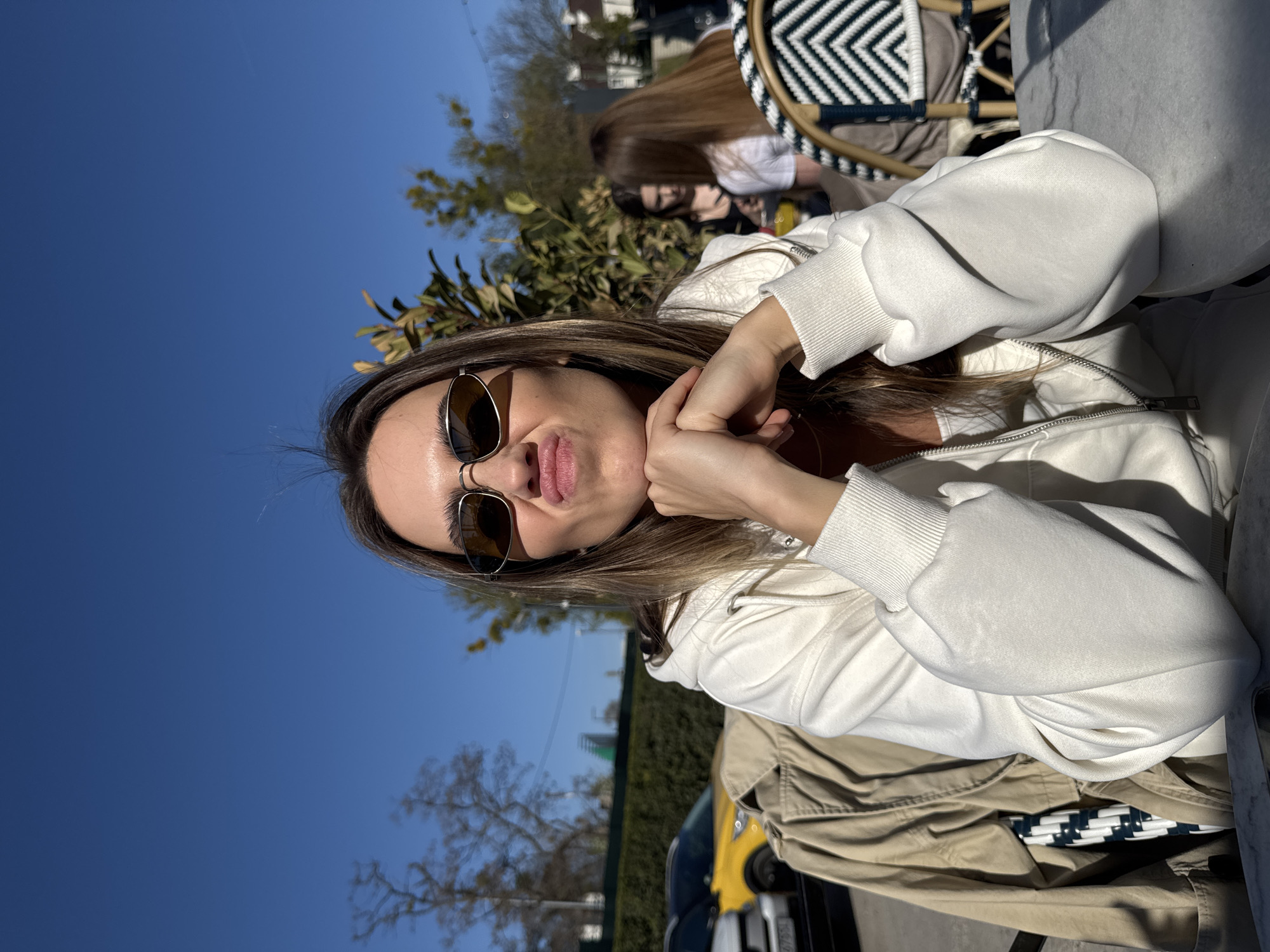 A woman with long, light brown hair and sunglasses is sitting at an outdoor table. She is wearing a white zip-up hoodie and has her hands under her chin, making a face for the camera. In the background, another person is sitting at a nearby table with a black and white woven chair. A tree, a green hedge, and a yellow car are also visible in the background, against a bright blue sky. Based on the surroundings, it could be a cafe in an urban area, potentially in the city of Warsaw, Poland.