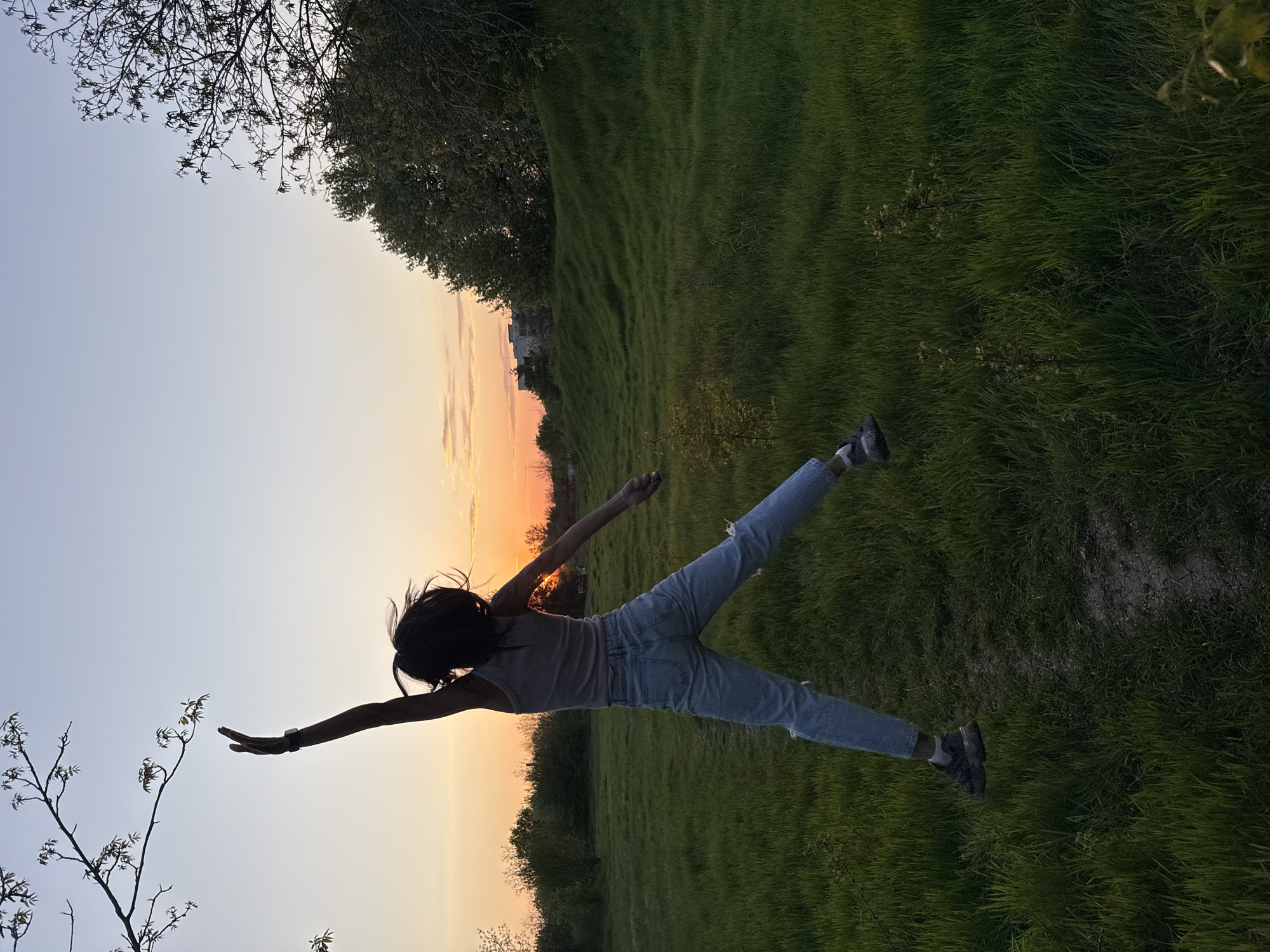 A woman is silhouetted against a sunset sky while laying horizontally on what appears to be a grassy field. Her arms are extended, and her legs are spread in a "flying" pose. She is wearing jeans and a light-colored top. The background consists of green grass, trees, and a sunset with orange and yellow hues. The location is potentially in the city of Pleven, Bulgaria.
