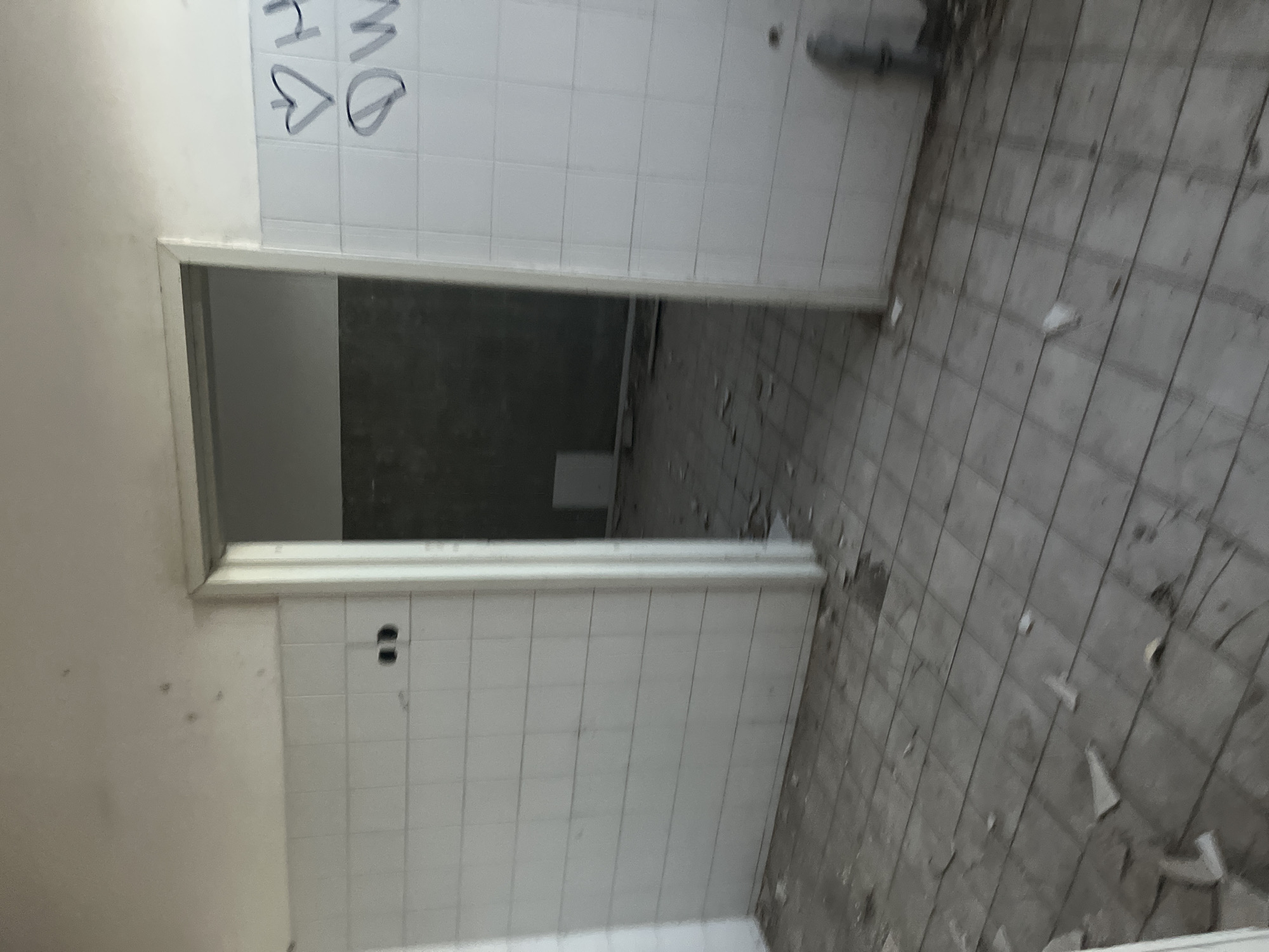 The image depicts an interior space, possibly an abandoned building. The walls are tiled with white, rectangular tiles, some of which are marked with graffiti. There's a doorway leading to a darker room, possibly a hallway. The floor is tiled in a different style, with grey, larger tiles showing signs of wear and damage, with scattered debris. The overall impression is that of neglect and disrepair. There are no people visible in the image. I do not have enough information to determine if the location is in Марковский, Russia.