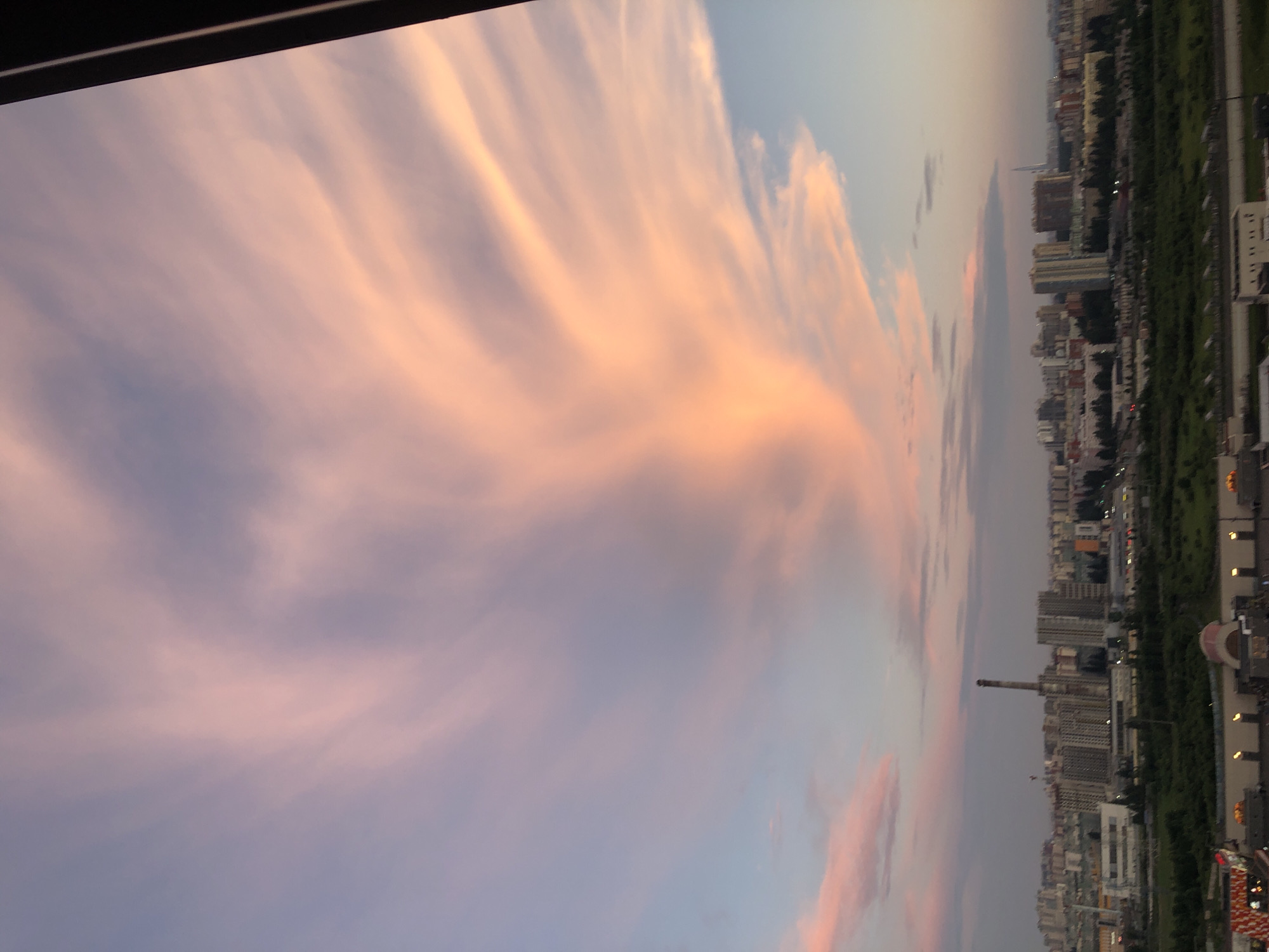 The image shows a wide-angle view of a city skyline at dusk, dominated by a dramatic sky filled with wispy, pink and orange clouds. In the foreground, there is a dark, vertical object on the right side of the frame, possibly a window frame or architectural element. The middle ground features a sprawling cityscape with numerous buildings of varying heights, including apartment blocks and what appears to be an industrial facility with a tall chimney. Trees and green spaces are visible between the buildings, suggesting parks or residential areas. The background is the sky, which transitions from a pale blue at the top to warmer hues of pink and orange near the horizon due to the setting sun. The overall impression is one of urban development set against a beautiful, atmospheric sky.