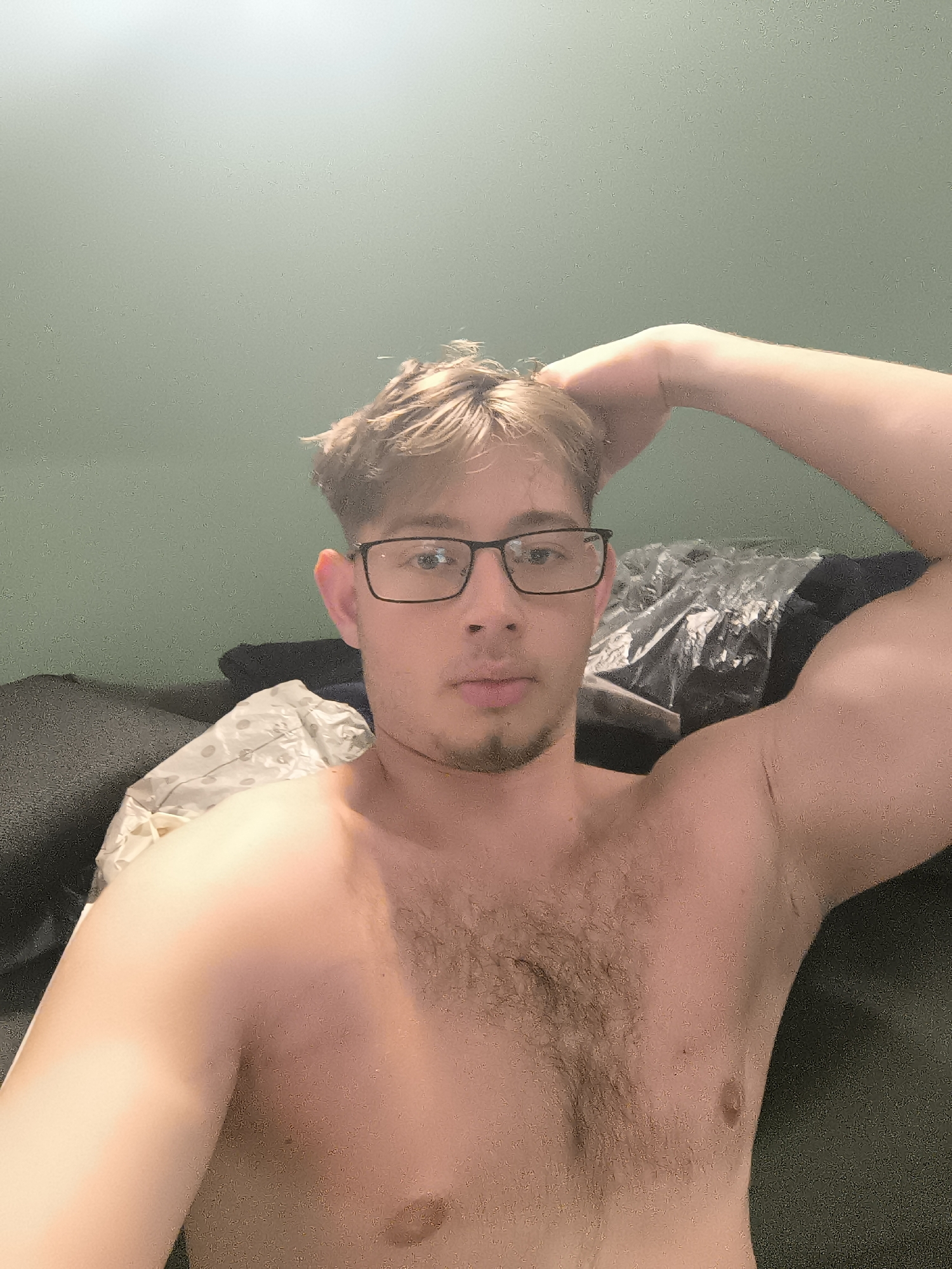 A shirtless man with light brown hair, wearing glasses, is reclining on a dark gray couch with a patterned white pillow behind him. He is touching his hair with his right hand and looking directly at the camera. The background is a plain green wall. The location and the city of Sofia, Bulgaria are not discernible from this image.