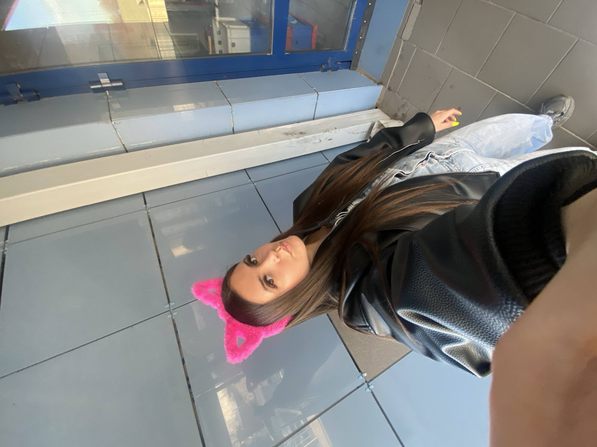 A young woman poses for a selfie in what appears to be an urban public space. She is wearing a black leather jacket, light blue jeans, and pink cat ears headband. The background consists of light blue tiled walls and grey tiled floors, with a window or display case visible behind her on the left. She is located in the city of Chaikovsky, Russia.