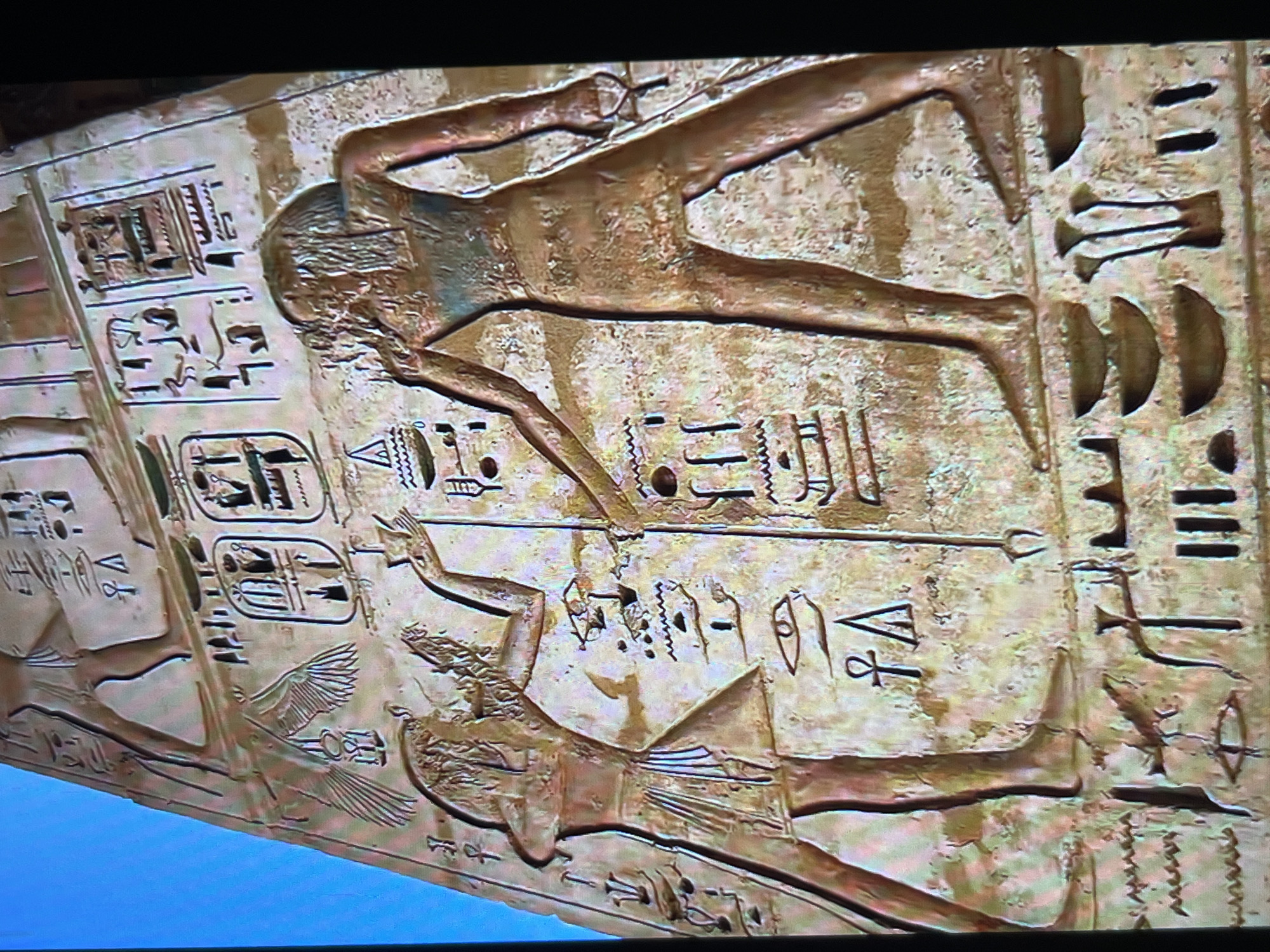 The image depicts a section of ancient Egyptian relief carving or hieroglyphs. Two figures are visible in profile. The upper figure is stretched out horizontally. The lower figure has wings. Hieroglyphic text accompanies and surrounds the figures. No information regarding the location of Tinaquillo, Venezuela, is present.