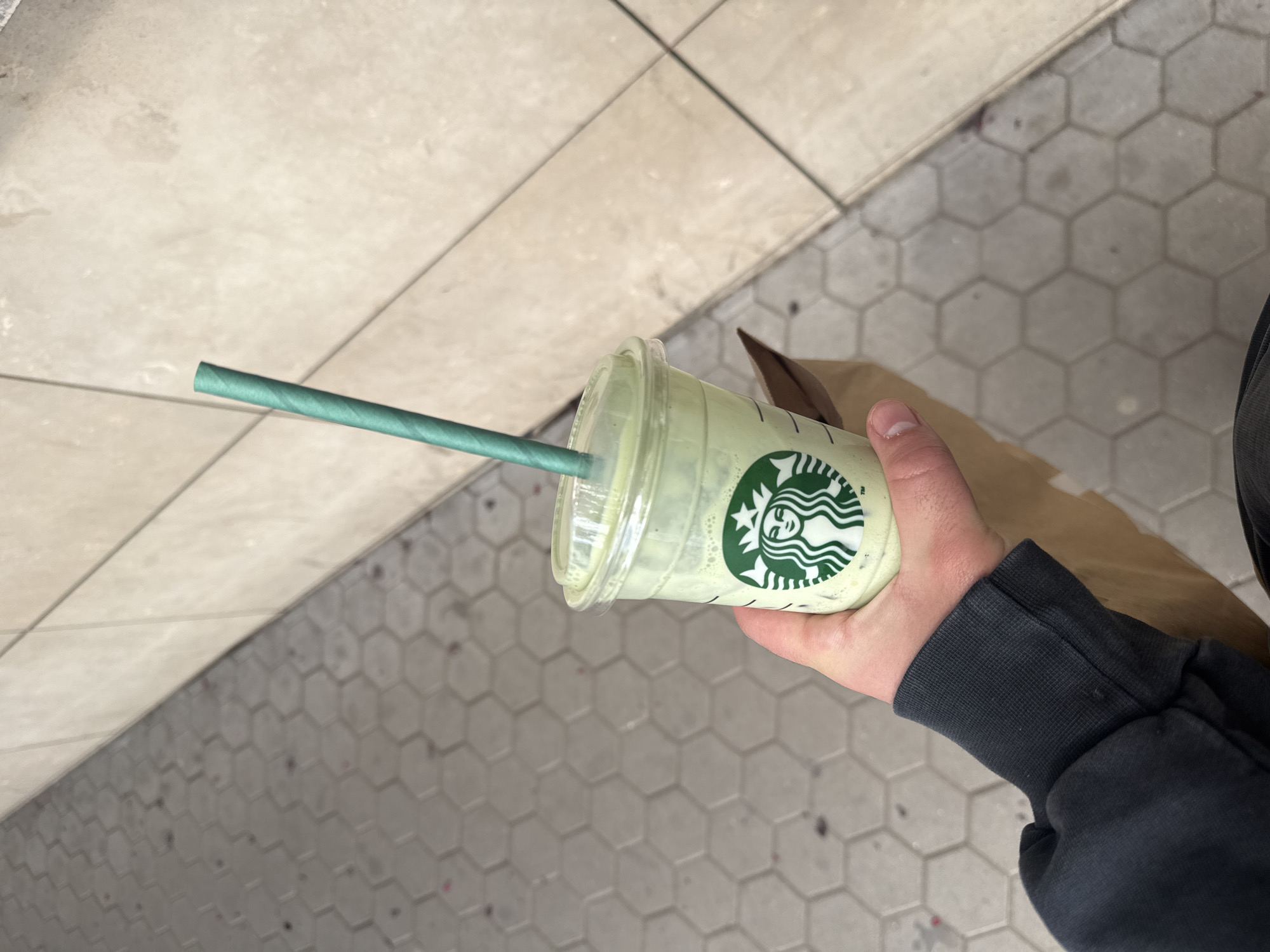 A person is holding a Starbucks drink, likely a matcha latte given its green color, with a green paper straw. The drink is in a clear plastic cup with the Starbucks logo visible. The person is wearing a dark grey or black long-sleeved shirt or jacket. The background shows a sidewalk or walkway with beige square tiles on one side and grey hexagon tiles on the other. Given the prevalence of Starbucks and the style of tiling, this image could conceivably be taken in a major city like Sofia, Bulgaria, but there is no specific evidence in the image to confirm the location.