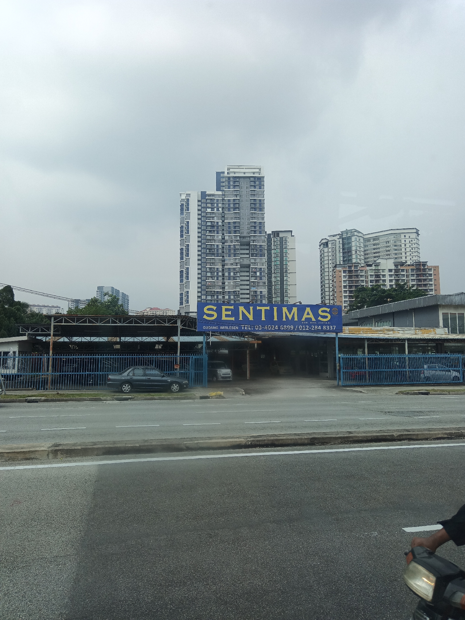The image shows a street view in Kuala Lumpur, Malaysia, under an overcast sky. In the foreground, a gray asphalt road with white lane markings is visible. To the right, the front of a motorcycle with a person's arm and hand holding the handlebars is partially captured. Behind the road, a blue metal fence encloses a property.
The property itself features a covered structure with a corrugated metal roof supported by metal posts, appearing to be an open-sided warehouse or workshop. A dark gray sedan is parked in front of this structure, with its rear facing the road. Another car is partially visible within the covered area.
Further back, a prominent building displays a large blue sign with yellow lettering that reads "SENTIMAS" and smaller text below including "SDN. BHD." and phone numbers. Adjacent to this is a modern, tall residential building with numerous windows. In the background, several other high-rise buildings, characteristic of a city skyline, are visible. Trees and utility poles are also present in the scene. The overall impression is that of a commercial or industrial area within a bustling city.