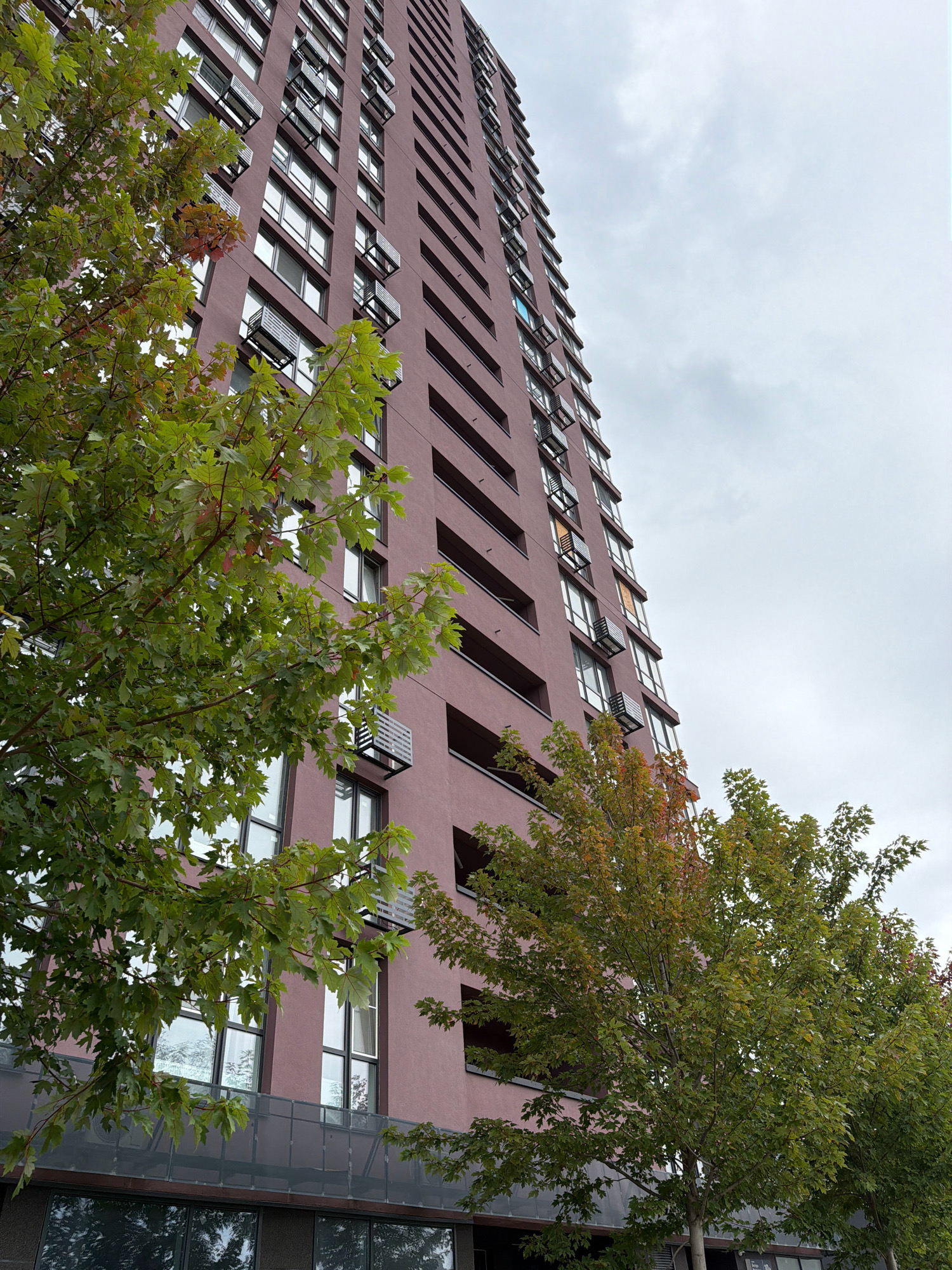 A low-angle shot captures the facade of a tall, reddish-brown apartment building in Kyiv, Ukraine. The building has numerous windows, some with open air conditioning units. In the foreground, leafy green trees partially obscure the lower floors of the building. The sky above is overcast and grey. The image appears to be taken on a street level.