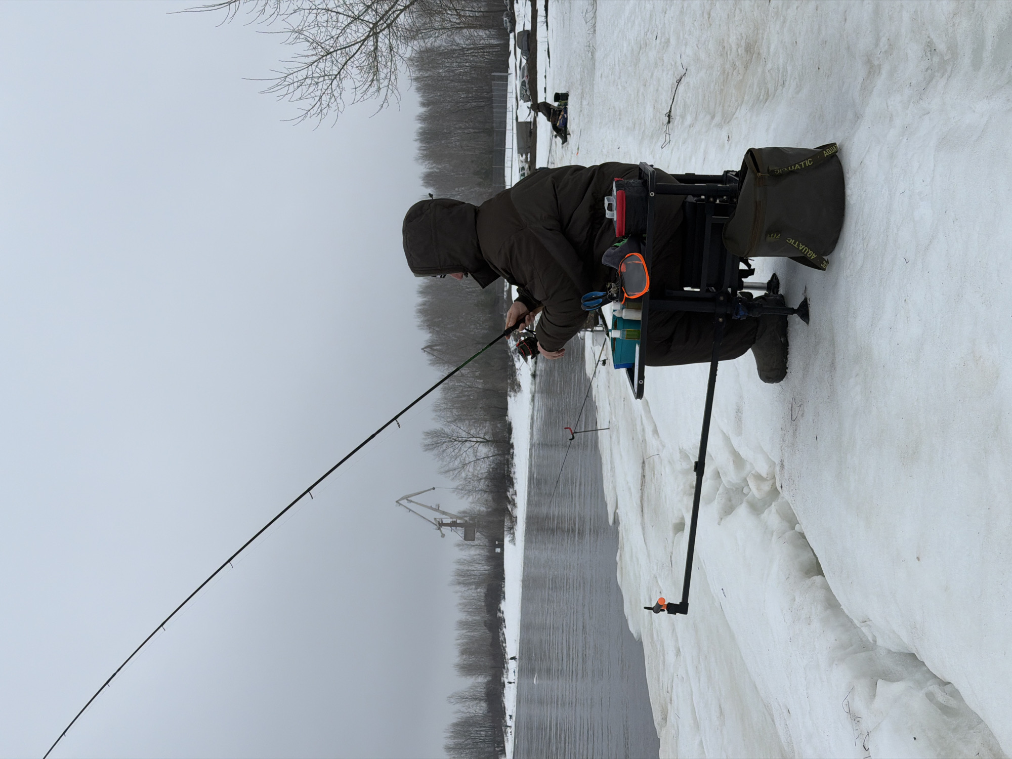 A man is fishing in the city of Chaikovsky, Russia. He is bundled in a hooded jacket and is seated on a platform chair with adjustable legs. The chair has attached storage units. He is holding a fishing rod, which is angled over the water. Snow covers the ground around the river. The sky is overcast. A crane can be seen in the distance across the water.