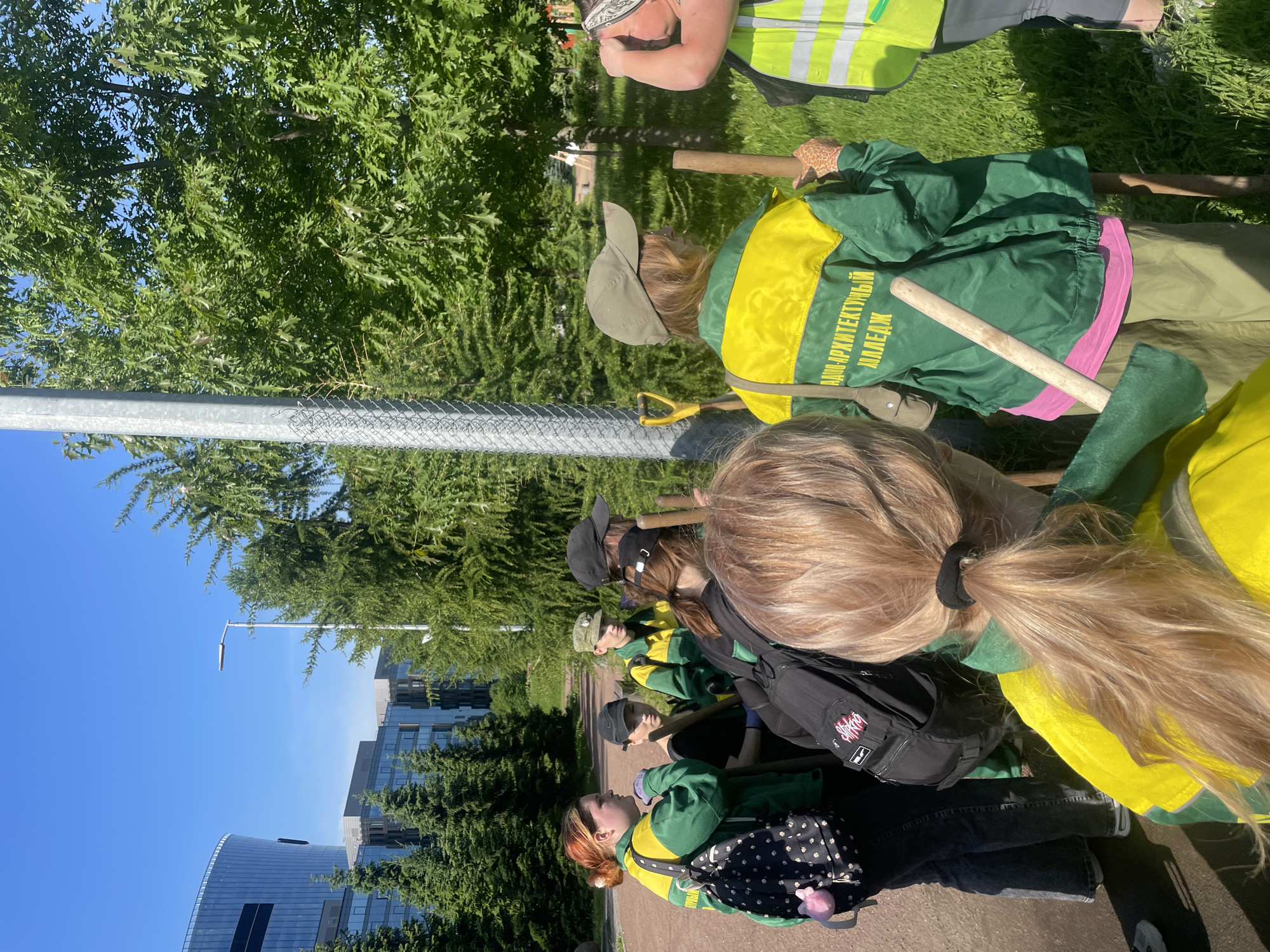 The image shows a group of young people wearing green and yellow work jackets, some with the text "АРХИТЕКТУРНЫЙ КОЛЛЕДЖ" (Architecture College) visible, gathered outdoors in a park-like setting with trees and green grass. They are holding shovels and appear to be engaged in some kind of outdoor activity, possibly planting or gardening. In the background, modern glass-fronted buildings and more trees are visible, suggesting an urban environment. The scene is lit by bright sunlight. One building in the background features a distinctive curved glass facade. The presence of the architecture college uniform indicates a possible connection to an educational institution. The environment appears to be well-maintained, with paved paths and manicured greenery. This location is likely part of the ITMO University campus or a similar modern development in Saint Petersburg, Russia.