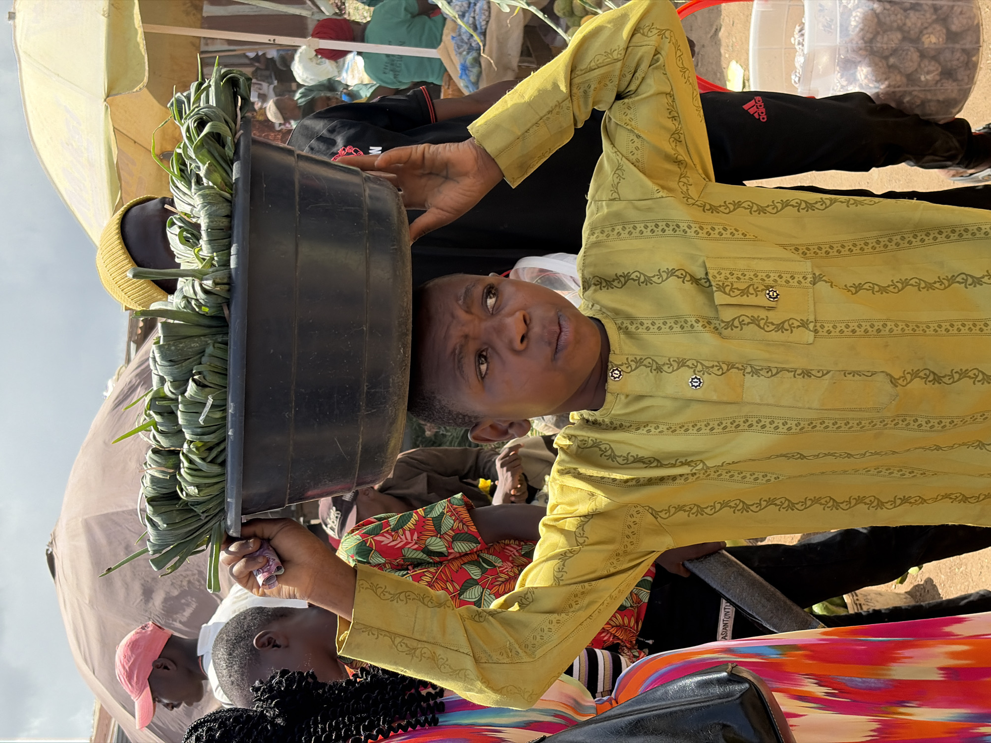 An eye-level shot captures a young person, likely a boy, carrying a large black basin filled with bundles of green leafy vegetables on his head. He looks directly at the camera with a neutral, inquisitive expression. He is wearing a light olive-green long-sleeved shirt adorned with intricate, dark green patterned borders and lines, featuring small, decorative buttons on the collar and shirt pocket. His right hand holds the rim of the basin to steady it, while his left arm is bent.
The setting appears to be a bustling outdoor market under bright daylight, indicated by the need for shade provided by a large, light yellow umbrella positioned to the left. The ground is dusty or sandy. Several other individuals are visible in the background and surrounding areas, some partially obscured. These include a person in a yellow knitted cap under the umbrella, another wearing a pink baseball cap, and various figures in colorful clothing. In the upper right background, a clear plastic container holds numerous small, dark, round objects, possibly nuts or seeds for sale. A person in the background wears black track pants with a red "Adidas" logo visible on the side. The scene depicts a common sight in market environments, where goods are often transported on heads, and vendors interact with their surroundings.