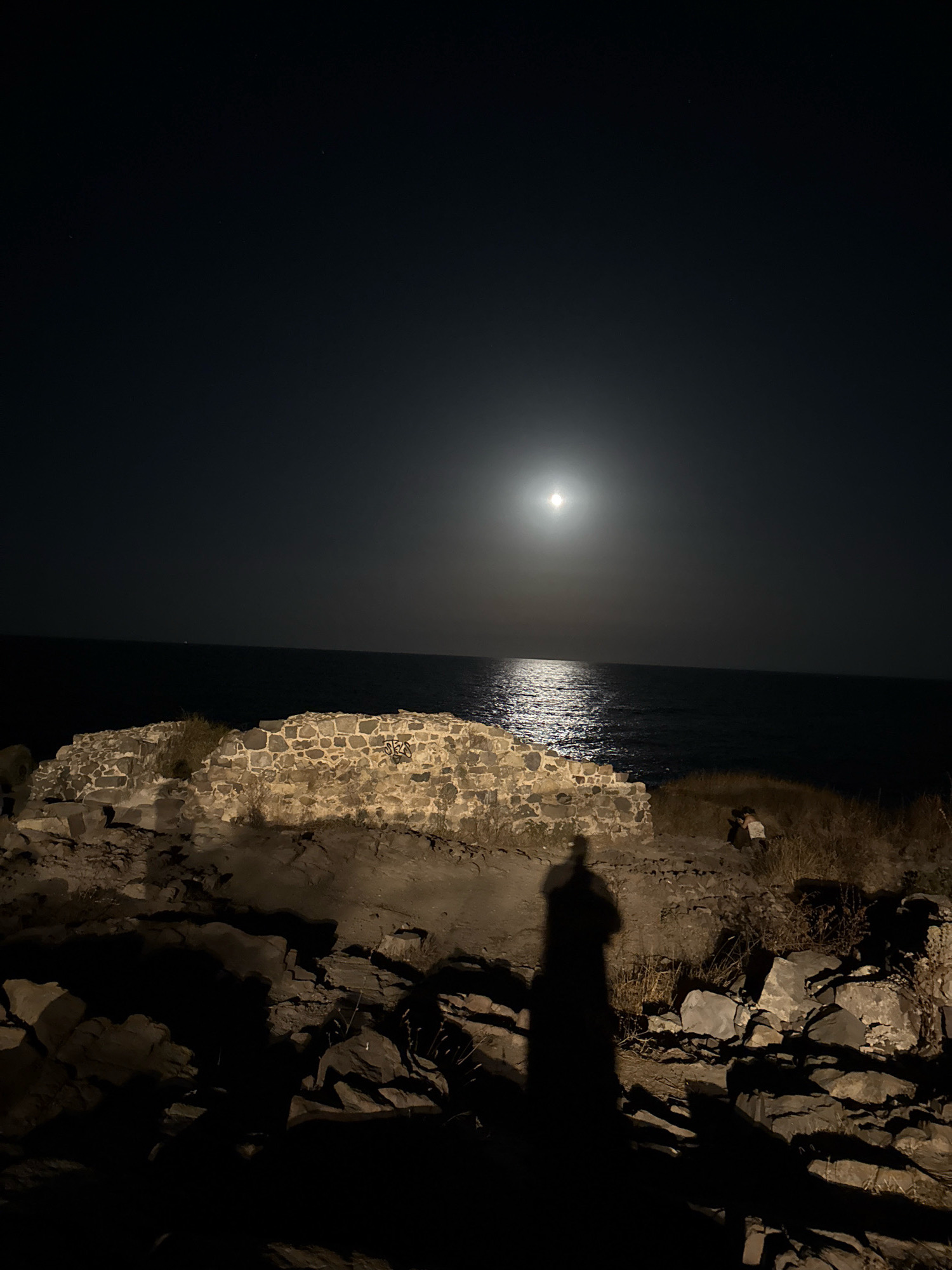 The image depicts a nighttime scene at the coastline in Sozopol, Bulgaria, under a full moon. A large, luminous moon is visible in the dark sky, casting a shimmering reflection on the surface of the calm sea. In the foreground, a silhouette of a person stands on a rocky outcrop, casting a long shadow. To the right and slightly behind the main shadow, another person is visible crouching, seemingly observing something or taking a photograph. In the middle ground, there are the remains of a stone structure, possibly an ancient wall or ruin, overgrown with sparse vegetation. The ground is covered in rocks and dry grass, indicating a natural, unmanicured coastal environment. The overall atmosphere is serene and somewhat mysterious, illuminated by the moonlight.