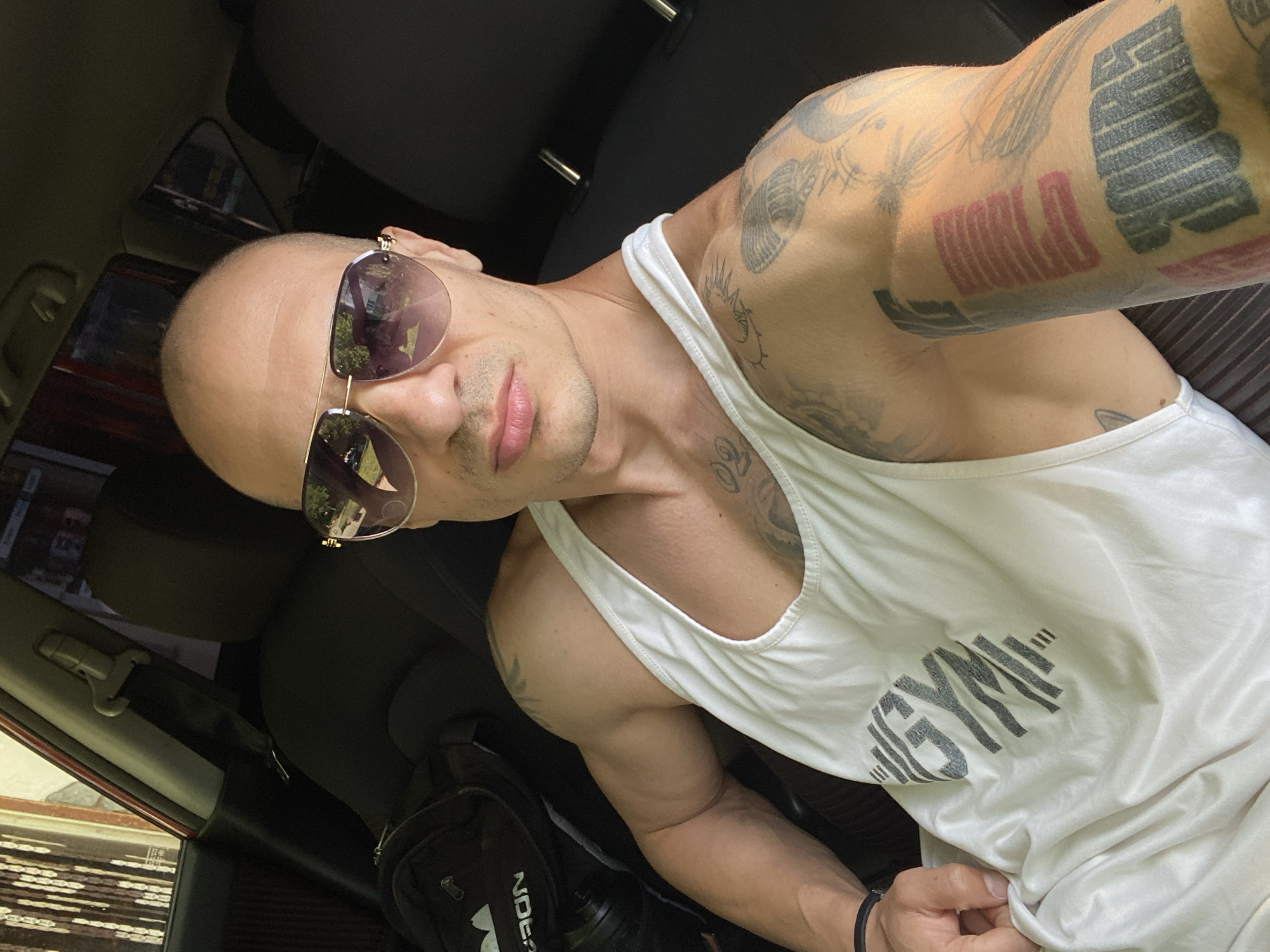 A tanned, bald man wearing gold-rimmed sunglasses and a white tank top with the word "GYM" printed on the lower right chest area, is pictured in the interior of a car. He is heavily tattooed on his left arm and chest. A black backpack is to his left. The location is not discernible as Pleven, Bulgaria, from this image. The man is taking a selfie, and the environment is dark inside the car.