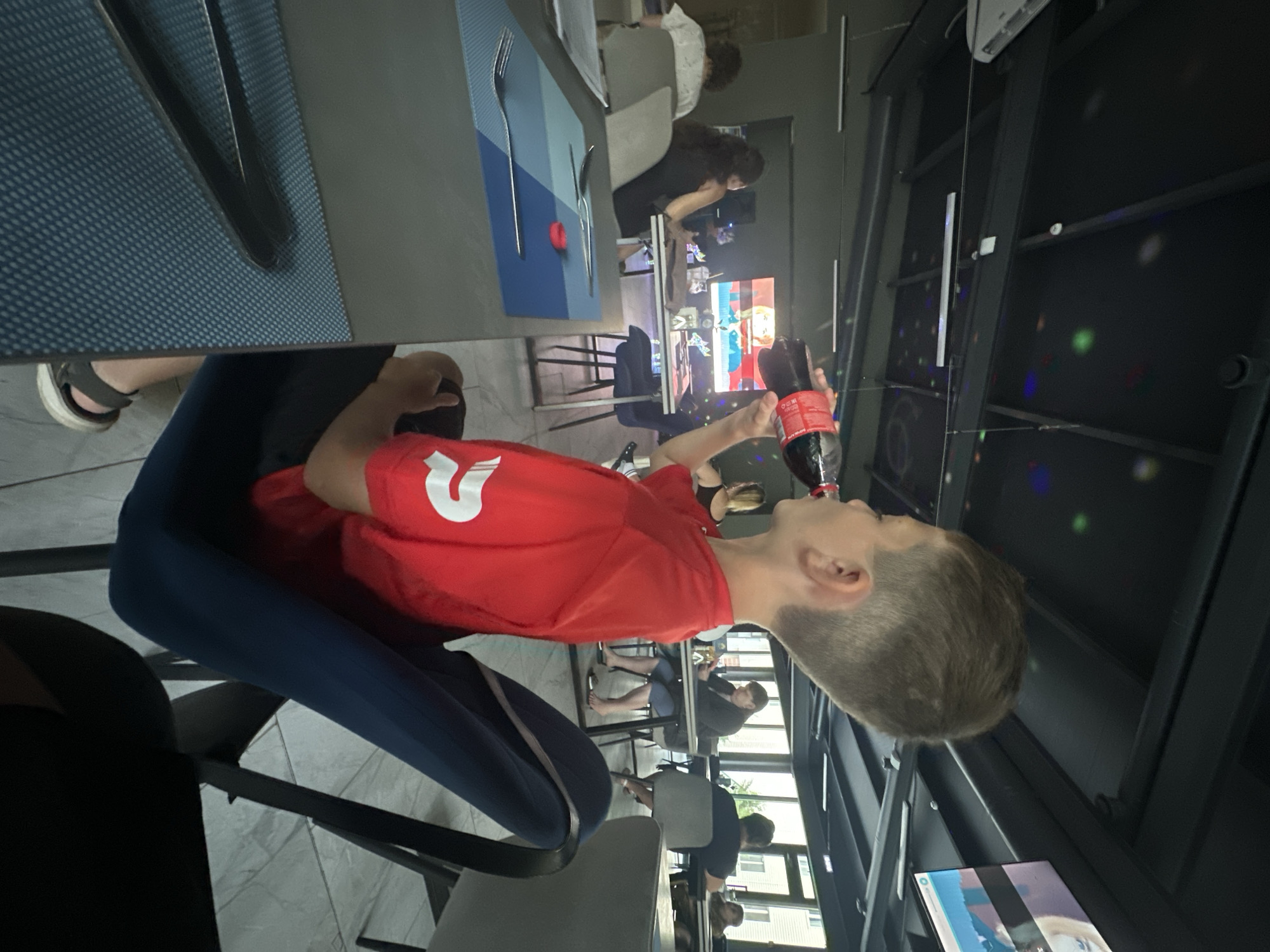 In this vertically oriented image taken in Danilovka (Altai Krai), Russia, a young person wearing a red t-shirt with a white logo is drinking from a bottle of Coca-Cola. The person is seated at a table in what appears to be a modern, indoor dining area. They are looking up and to the side, with their arm bent as they bring the bottle to their mouth. The red t-shirt has a prominent white "2" on the chest. The person is wearing dark blue leggings and sandals.
The setting features a dark-colored table with a textured blue placemat and silverware. In the background, other patrons are visible, some seated and some standing. One person in the background is wearing a white shirt and another is wearing a black dress. There is a screen in the background displaying what looks like a cartoon or animation. The ceiling has a black, textured appearance with several small, colored lights that resemble a disco ball effect. The overall ambiance suggests a casual gathering or meal. The lighting is somewhat dim, with artificial lights contributing to the atmosphere. The image seems to have been taken during the daytime, but the interior lighting is prominent.