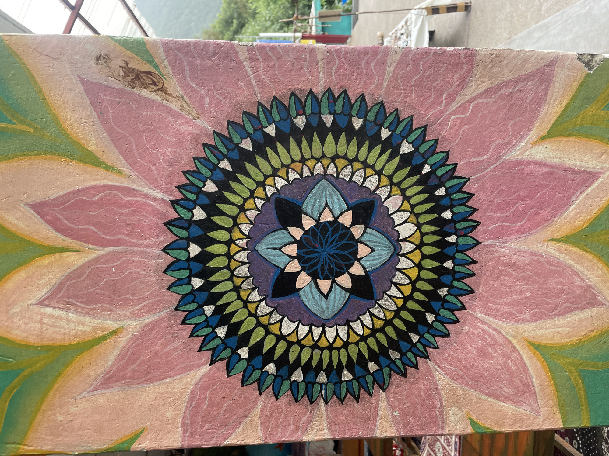 The image shows a close-up of a colorful, intricate painting of a lotus flower on a textured, rectangular surface, likely a wall or pillar. The painting features multiple layers of petals in shades of pink, yellow, and green, radiating outwards from a central, detailed mandala design. The mandala itself is composed of concentric circles with repeating geometric patterns in blue, green, black, white, and yellow. The surface of the painting appears slightly worn and textured, with visible brushstrokes and some minor imperfections. In the background, out of focus, there is a hint of an outdoor environment with some structures and greenery, suggesting it is located in an open area. There are no people or animals visible in the frame. This appears to be a decorative artwork located in Haini, India.
