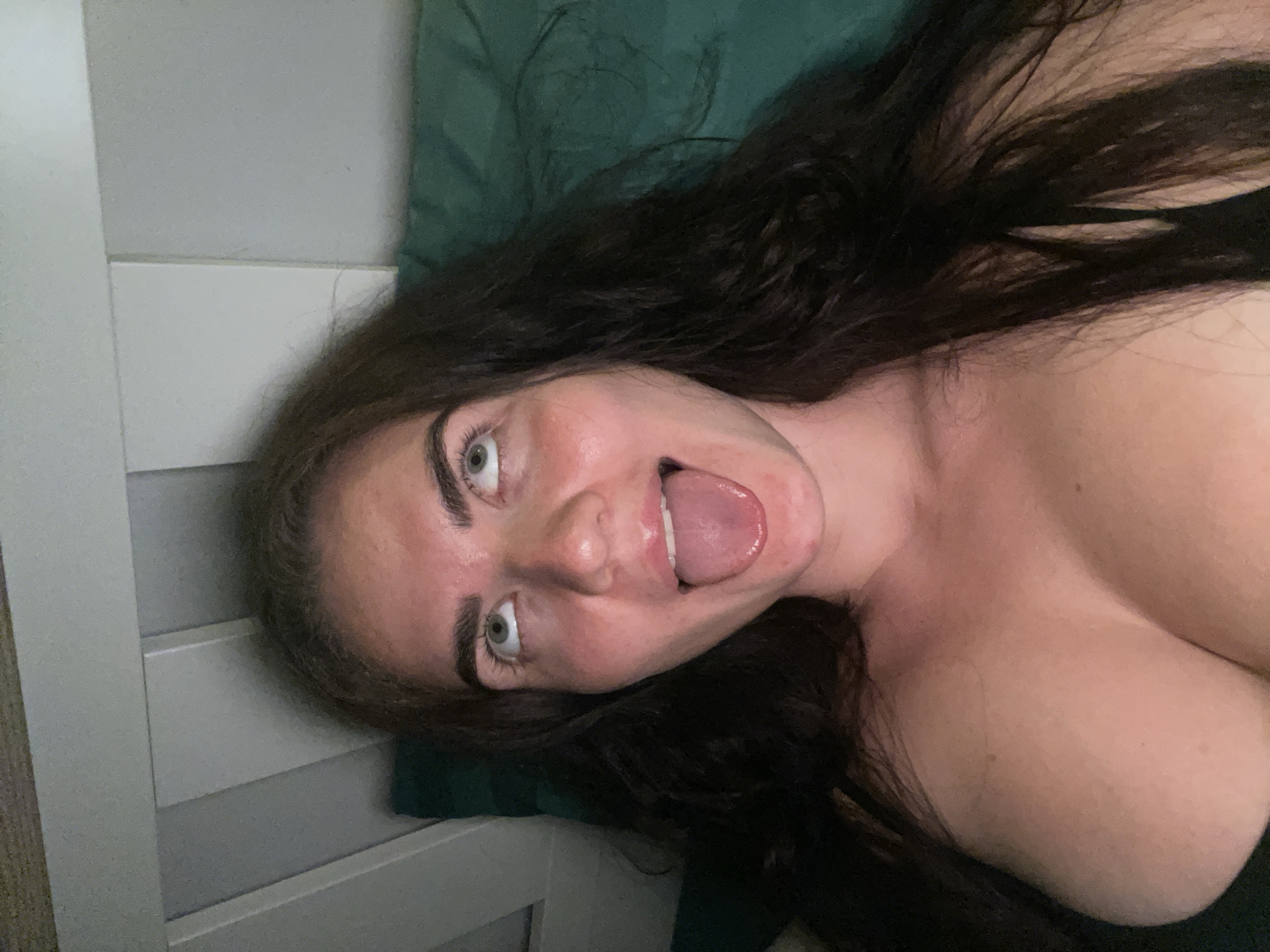 The image is a close-up, over-the-shoulder shot of a woman with long, dark hair. She is looking upwards with her tongue sticking out. She is wearing a dark top that exposes her cleavage. The background appears to be a bed with a dark green pillow and a white wooden headboard. The lighting is dim. The image does not contain any information about the city of Мосметаллоконструкция Онт, Russia.