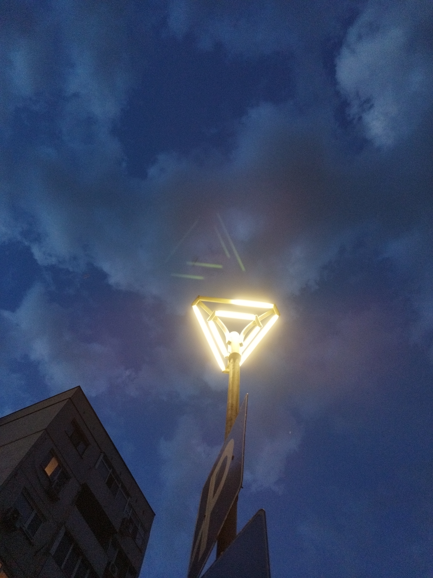 An upward shot shows a street lamp against a blue, cloudy evening sky. The street lamp has a triangular design and is illuminated with a warm, yellow light. Below the lamp, there is a sign. Part of a building can be seen to the left side of the image. The photo may have been taken in Sofia, Bulgaria. No people are visible in the photo.