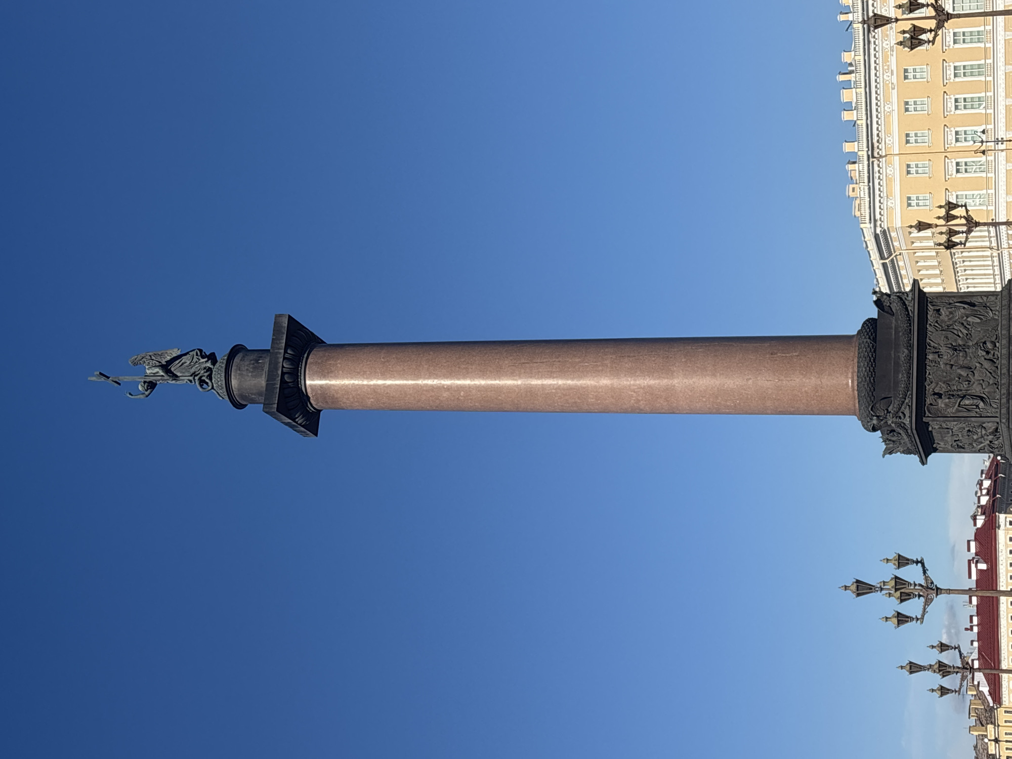 The image displays a full-height view of the Alexander Column, located in Palace Square, Saint Petersburg, Russia. The column itself is a tall, monolithic shaft of polished red granite, rising vertically against a clear, vibrant blue sky. It is topped by a dark-bronze capital which supports a sculpture of an angel. The angel, depicted with outstretched wings and flowing garments, holds an upright cross in its raised left hand while its right hand points downwards.
The column rests on a large, square, dark-bronze pedestal. This pedestal is intricately decorated with bas-reliefs that appear to depict allegorical figures and military trophies. At the base of the column, where it meets the pedestal, a serpent motif is visible, potentially representing an Ouroboros.
In the background, on the right side of the image, a large, light-yellow neoclassical building with multiple white-framed windows and white architectural trim is visible, consistent with the General Staff Building. On the lower left and bottom center, several ornate, multi-lamp streetlights with a historical design are partially visible. Further to the left and behind these streetlights, parts of other yellow buildings and a building with a red roof can be seen. No individuals or discernible actions are present in the depicted scene, which is brightly illuminated by natural light.