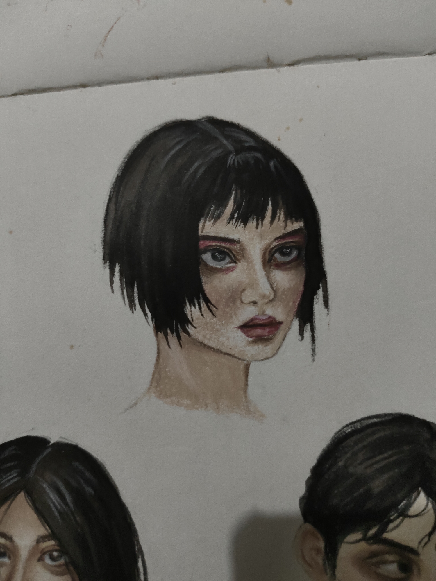 The image displays a watercolor or mixed media drawing of three partial human portraits on a piece of white paper. The central and most prominent subject is a portrait of a person with pale skin, dark, choppy bob-length hair, and short bangs. The subject's head is tilted slightly to the right, with direct eye contact. Heavy red eyeshadow is applied to both upper and lower eyelids, and there are dark, reddish-brown shadows beneath the eyes. The lips are closed and colored a muted reddish-brown. The neck and upper chest are depicted, fading into the uncolored paper.
Below and to the left of the central figure, a partial portrait shows the eyes and the top of the head of another person with dark hair and a fair complexion. Below and to the right, a third partial portrait reveals a side profile including an ear, dark hair, and a partially visible eye.
The background is a plain white paper surface exhibiting minor imperfections, including several small, light brown spots or smudges scattered across it. A distinct horizontal crease runs across the top portion of the paper, above the central portrait. The overall impression is a page from a sketchbook or a collection of studies. No external environmental or location-specific details are visible within the image.
