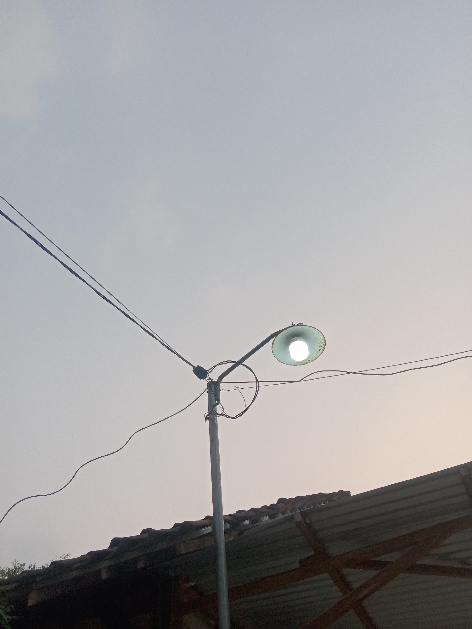 An outdoor, low-angle shot captures a streetlamp with a glowing bulb against a hazy, pale sky. The lamp is attached to a metal pole, with visible wires extending from it. A section of a tiled roof and some dark structures are visible in the lower portion of the frame, suggesting an urban or semi-urban environment. The image appears to be taken during the daytime, possibly at dusk or dawn due to the soft lighting. The location is identified as Jumput, Indonesia.