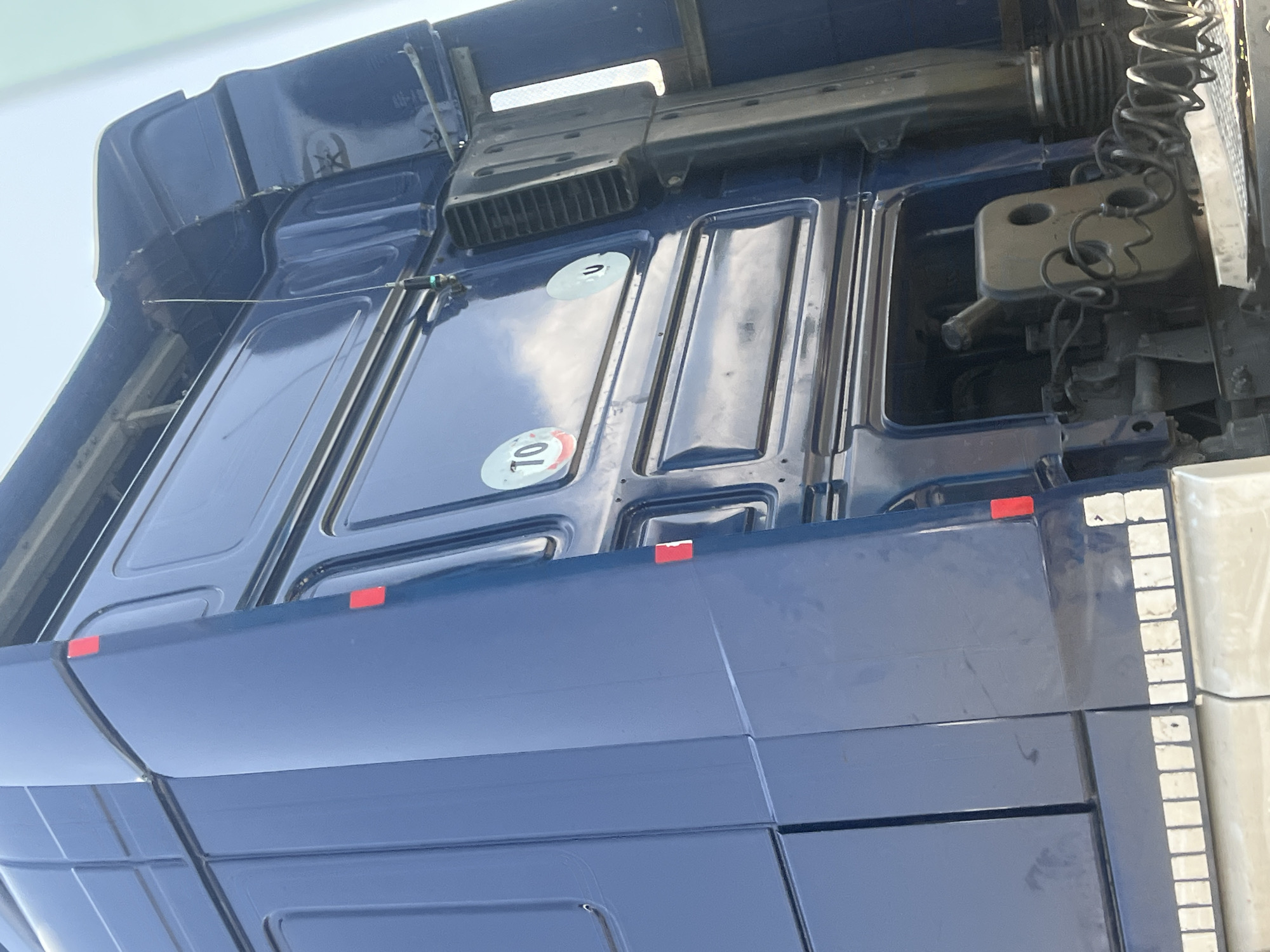 The image displays the rear-right section of a dark blue semi-truck tractor unit, captured from a low angle under a clear, bright sky in the city of Balanga, Nigeria. The truck cab is equipped with vertical aerodynamic panels on its rear, each featuring three small, rectangular red reflective markers. Two circular decals are affixed to the cab's rear paneling: one clearly shows "70" in red text on a white background with a red border, while the other, partially visible, displays a faded "U" or similar character. A slender, metallic whip antenna extends vertically from the upper-right rear section of the cab. Below the cab, a robust, dark-colored fifth-wheel coupling mechanism is visible, accompanied by several coiled air and electrical lines positioned ready for trailer connection. The lower portion of the chassis frame, appearing in a lighter grey tone, features a horizontal strip of rectangular white reflective markers that show signs of wear. Integrated ventilation grilles are present on the rear structure of the cab above the fifth-wheel assembly. No individuals are visible within the frame.