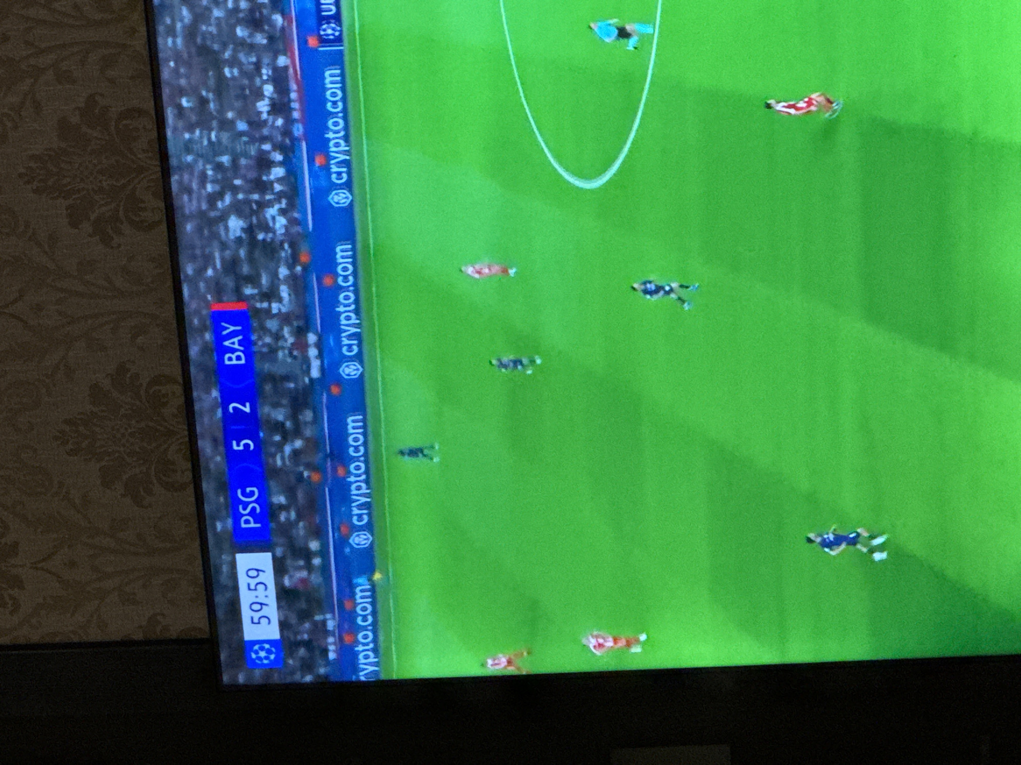 A television screen displays a football (soccer) match in progress, captured at an oblique angle. The screen shows a vibrant green pitch with several players on the field. Players are visible in different colored kits: red and white, blue and white, a darker blue/black, and one player (likely a goalkeeper) in light blue or teal near a goal area. A white semi-circular arc, part of the penalty box, is also visible.
Along the left side of the screen, a scoreboard overlay provides game information. It shows "PSG 5" and "BAY 2", indicating a score of 5-2 between Paris Saint-Germain (PSG) and Bayern Munich (BAY). Below the team names, the game clock reads "59:59," signifying that the match is nearing the end of the 60th minute. A UEFA Champions League star-ball logo is visible next to the timer.
Multiple digital advertisement banners for "crypto.com" are displayed along the edge of the pitch, stretching vertically down the left side of the screen. The background behind the television screen features a light brown wallpaper with an intricate, darker brown floral or damask pattern. The overall image quality suggests it was taken with a camera pointing at a television screen, leading to some minor blur and distortion.