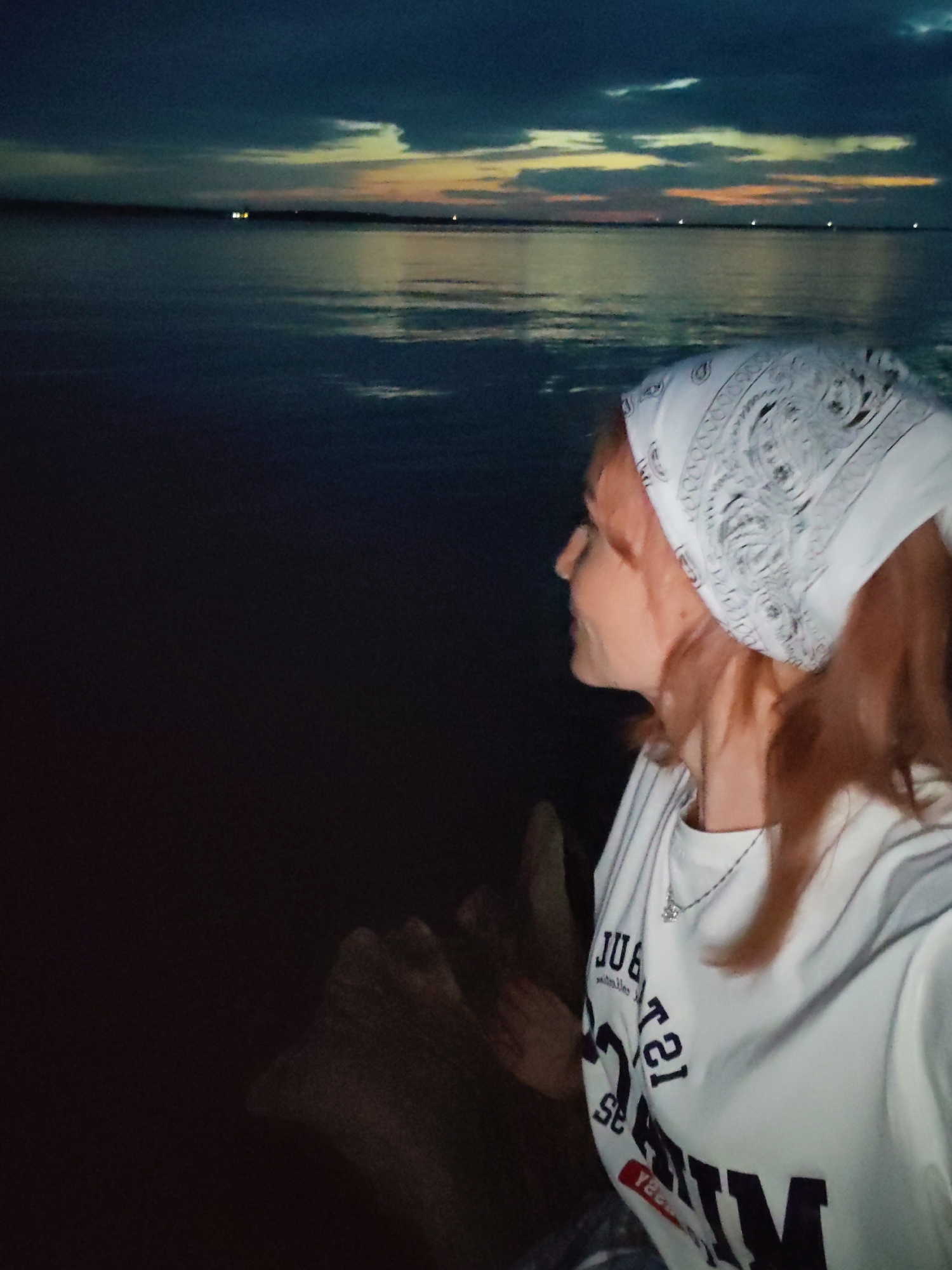 A young woman with red hair and a white bandana on her head is seated facing left, looking out at a body of water. The sky is dark with hints of orange and yellow from a sunset or sunrise. The water is dark and reflective, with some distant lights visible on the horizon. The woman is wearing a white t-shirt with text on it and a necklace. She is positioned in the foreground to the right of the image, with the water and sky occupying the rest of the frame. The image appears to have been taken outdoors at dusk or dawn. The location is likely near a body of water in Chaikovsky, Russia, given the context of the request.
