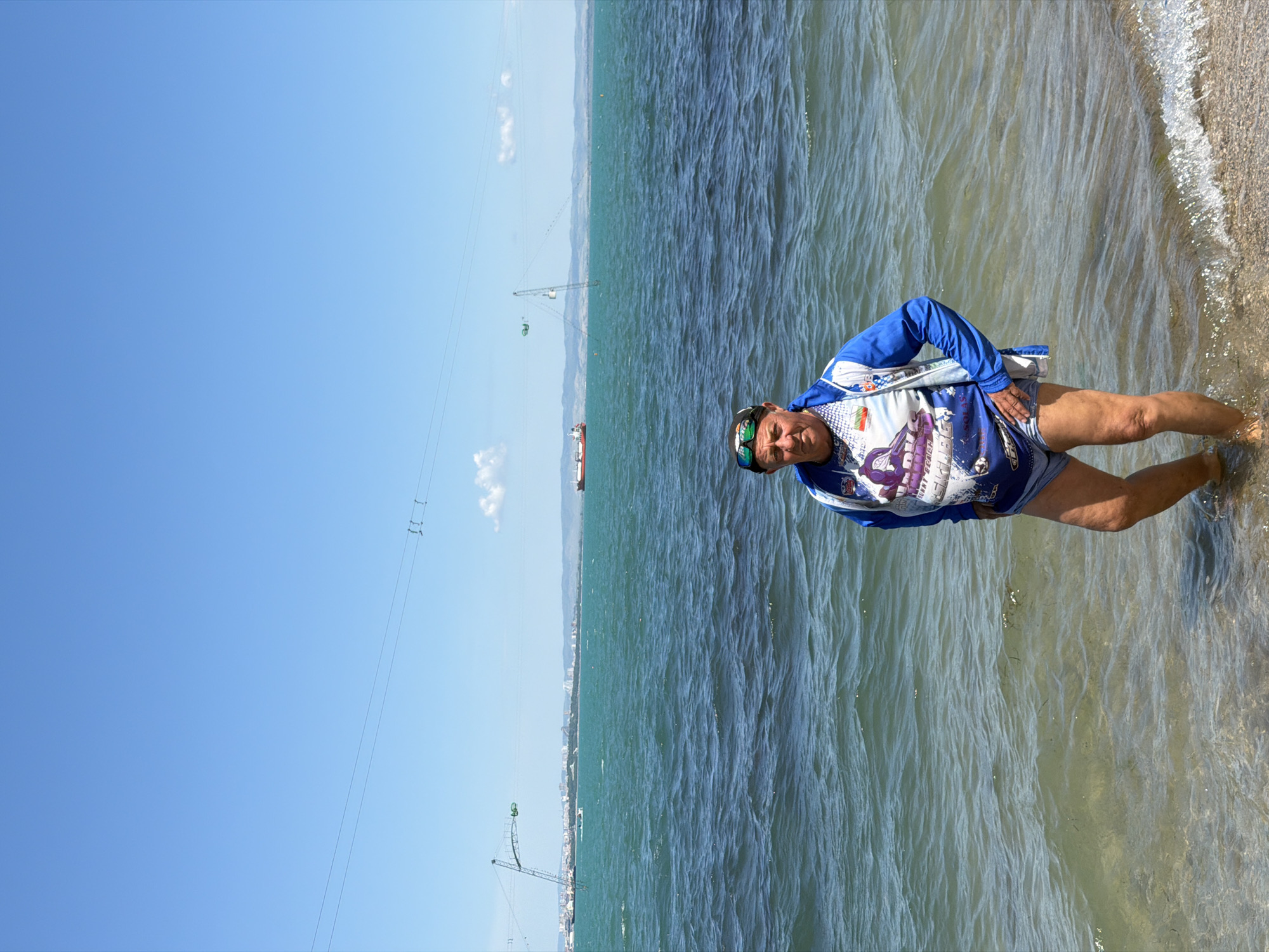 The image shows a middle-aged man standing in shallow water on a beach. He is wearing a blue and white long-sleeved shirt with cartoon graphics, blue shorts, a black hat with green accents, and sunglasses. The man is standing with one hand on his hip and looking at the camera. The water is clear and shallow near the shore, with small waves lapping at the beach. The beach consists of sand and small pebbles. In the background, there is a view of the sea, with a coastline visible in the distance. A ship is visible in the distance on the water. Power lines stretch across the sky from the left side of the image. Cranes are visible near the beach, and the image is taken in Burgas, Bulgaria, based on the location. The sky is a clear blue.
