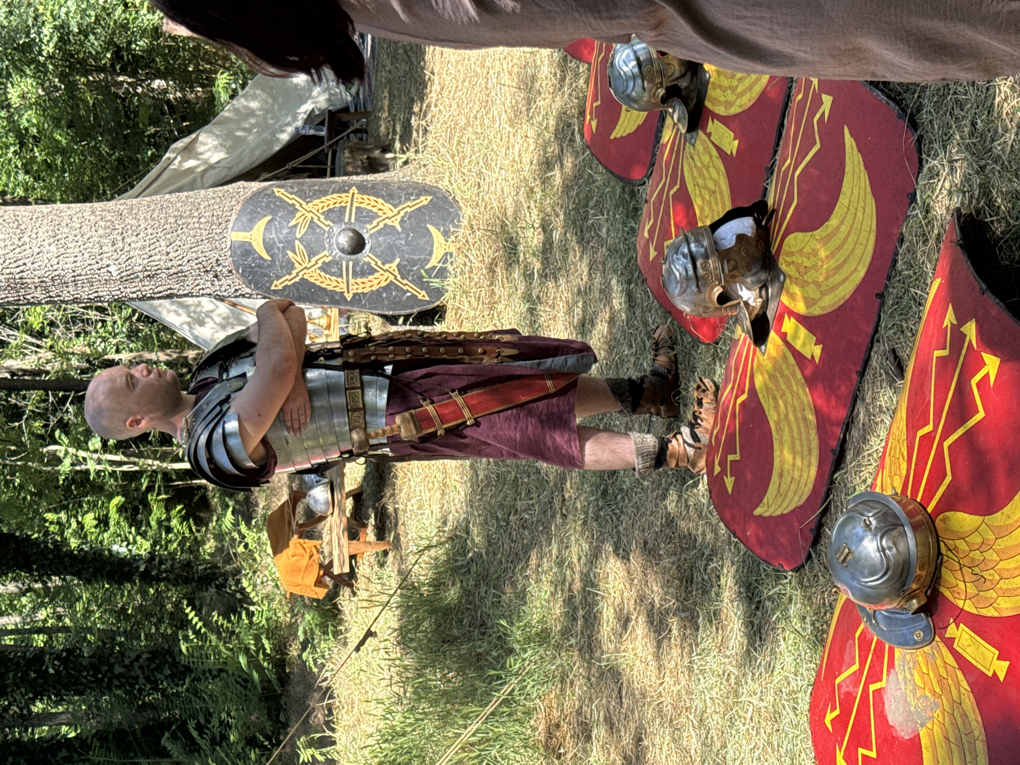 In a sun-dappled forest clearing, possibly within the bounds of Plovdiv, Bulgaria, a reenactor dressed as a Roman legionary stands with his arms crossed, gazing to his right. He wears a segmented metal cuirass, a burgundy tunic, and leather sandals with woolen leg wrappings. A scabbard containing a dagger is attached to his belt. In the foreground, several Roman shields, painted red with yellow eagle motifs, are laid on the ground, each adorned with a metal helmet. To the legionary's right, a circular Roman shield featuring a gold wreath and stylized crossed swords is propped against a large tree. Behind the legionary, a canvas tent and a wooden table with an orange cloth are visible, suggesting a temporary encampment or historical demonstration. The environment is characterized by lush green foliage and dry grass, indicative of an outdoor setting.
