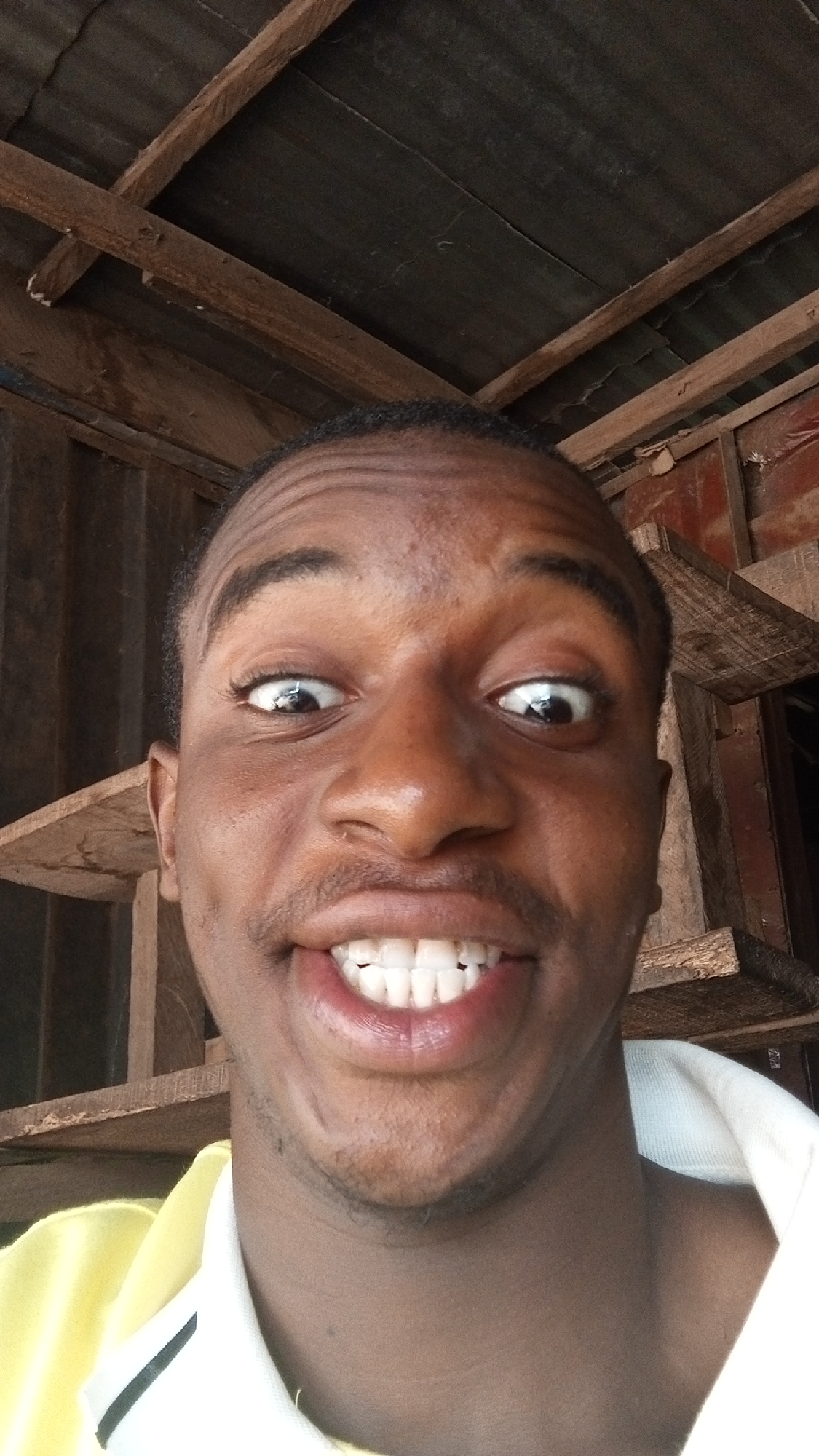 A close-up selfie of a young Black man with a wide smile, showing his teeth, and eyes slightly crossed. He has short hair and appears to be indoors under a wooden structure, possibly a roof or ceiling. Shelving is visible behind and to the side of him. The location is likely Onitsha, Nigeria. The man is wearing a collared shirt.