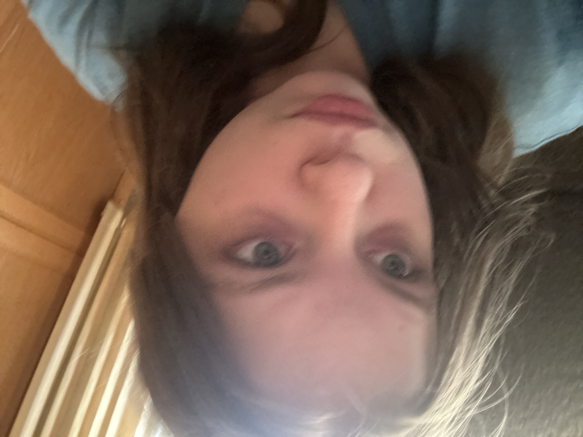 A selfie-style image shows a person with light skin, possibly female, with brown and blonde hair. The person is wearing a blue garment, possibly a shirt. The background features a wooden paneled wall and some light-colored curtains. No information suggests the location is Moscow, Russia.