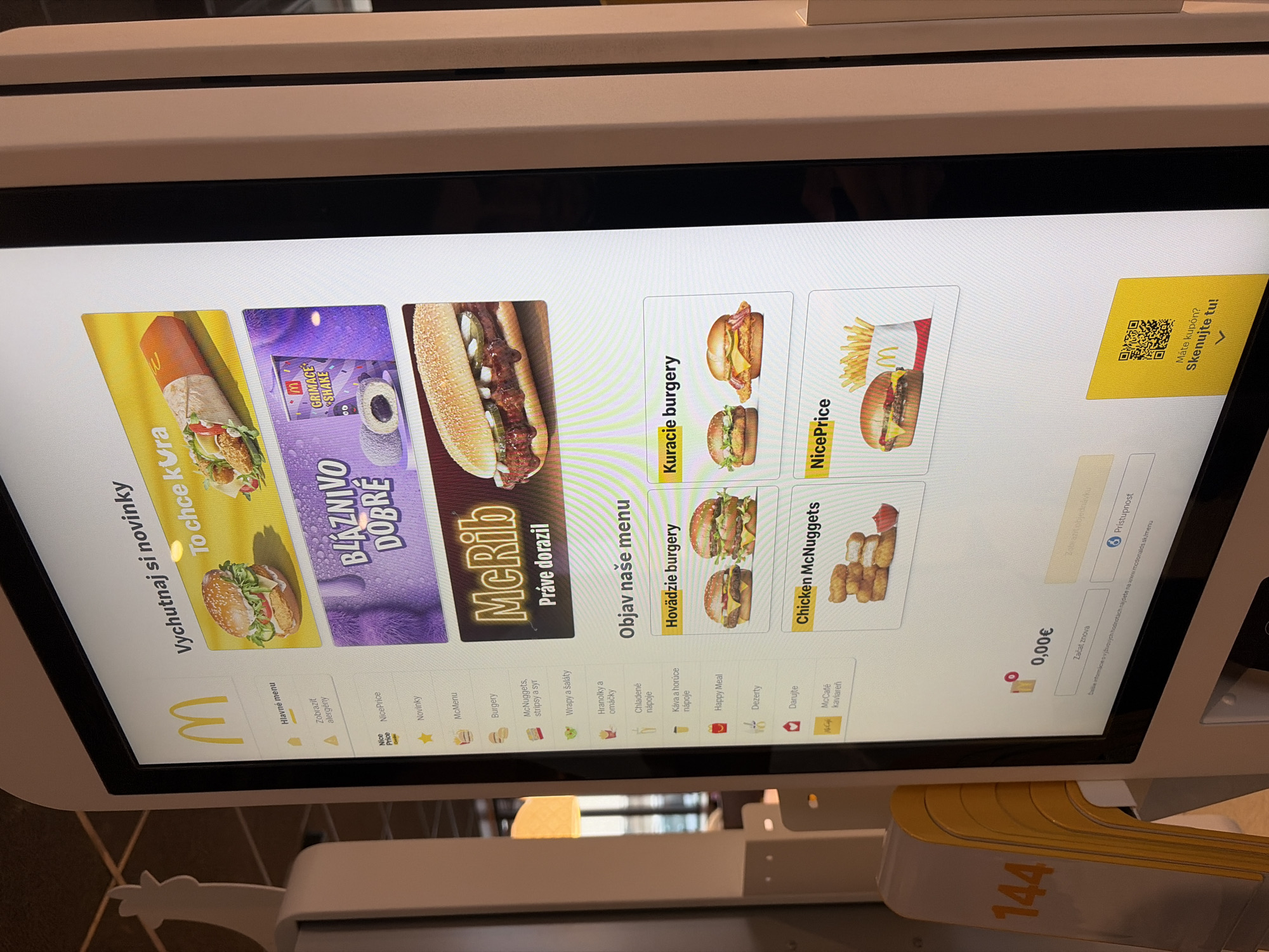 The image shows a close-up of a McDonald's self-order kiosk screen in Nitra, Slovakia. The screen displays various menu items, including "Vychutnaj si novinky" (Enjoy our new items), featuring a wrap and a burger, and "McRib Práve dorazil" (McRib just arrived). Below these promotional items, there is a section titled "Objav naše menu" (Discover our menu) which categorizes food items such as "Hovädzie burgery" (Beef burgers), "Kuracie burgery" (Chicken burgers), and "Chicken McNuggets". Further down, there's a "NicePrice" section with a burger and fries. The bottom of the screen shows a total cost of "0,00€" (0.00 Euros), a button to "Začať znova" (Start over), and a QR code with the text "Máte kupón? Sklenujte tu!" (Do you have a coupon? Scan here!). A yellow stand with the number "144" printed on it is visible on the left side of the image, partially obscuring the kiosk. The environment appears to be indoors, likely within a McDonald's restaurant.