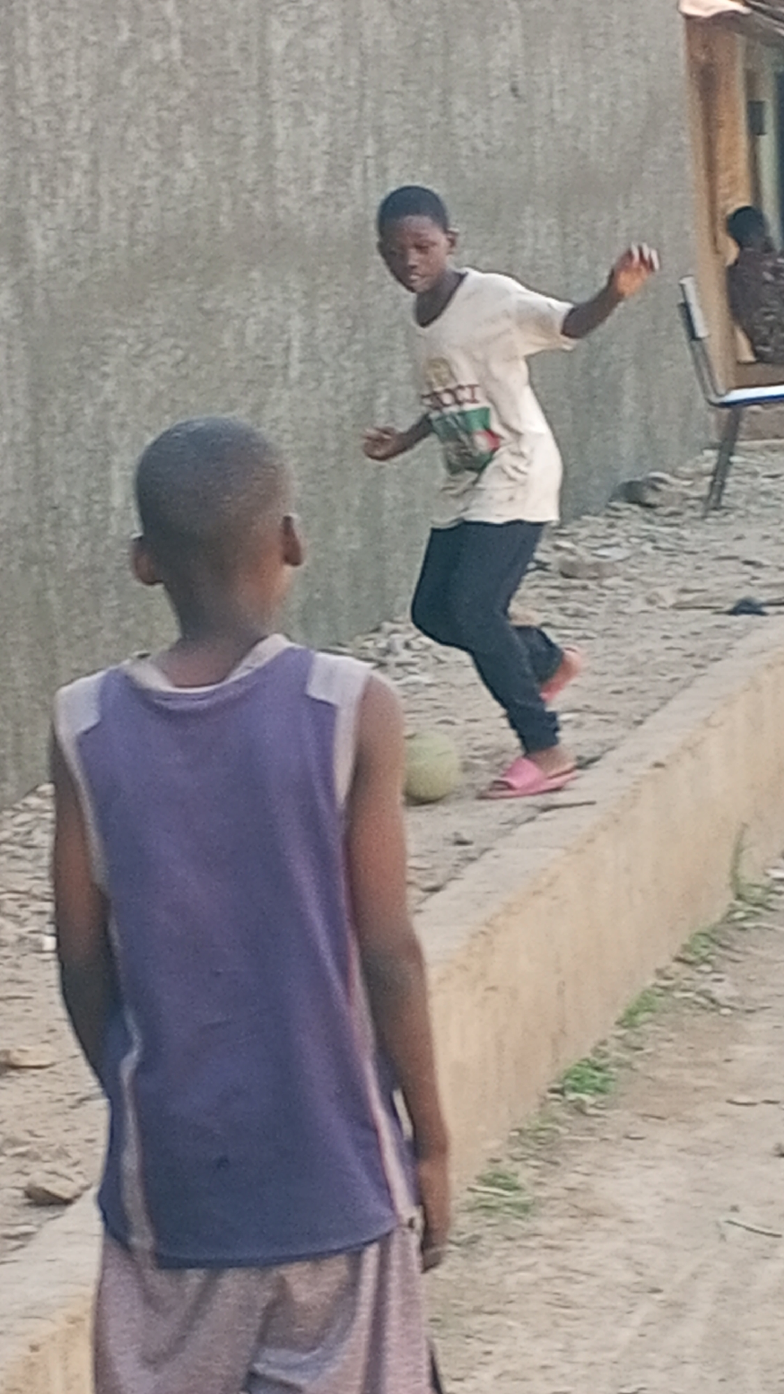Two boys are playing. One boy, wearing a purple tank top and grey shorts, is standing with his back to the camera. The other boy, wearing a white t-shirt, dark pants, and pink sandals, is kicking a ball on a low concrete ledge. The background shows a cement wall, a building with a chair outside, and a sandy ground. The scene is in Sabon Tasha, Nigeria.