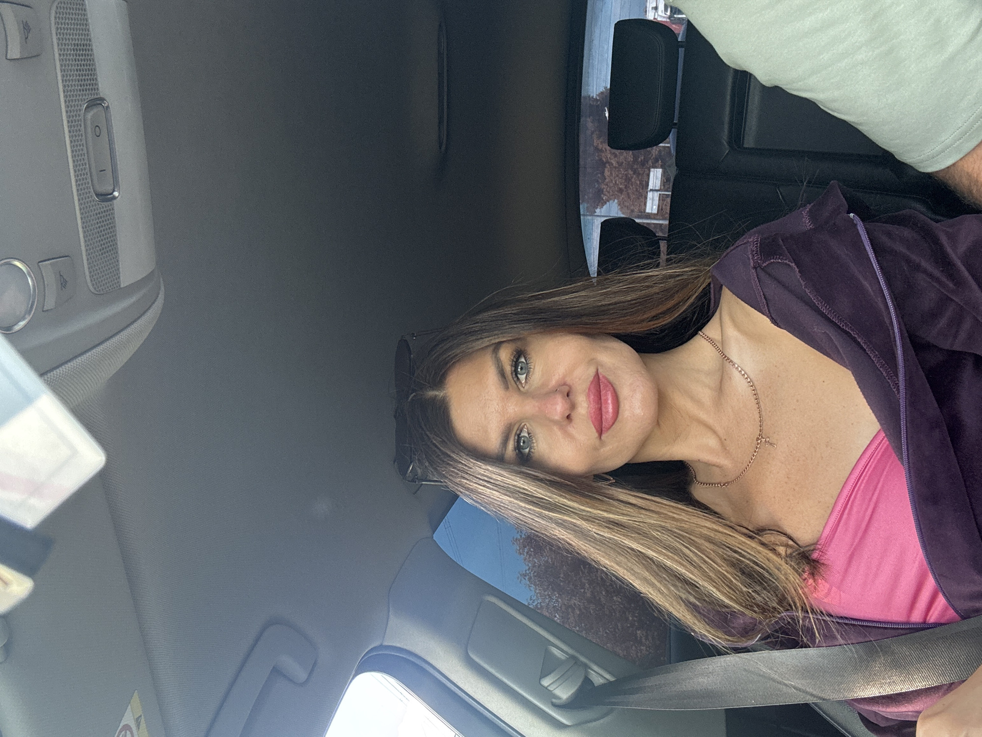 The image displays a woman seated in the driver's seat of a car, taking a selfie in the city of Chaikovsky, Russia, during daylight hours. The woman, appearing to be in her 30s-40s, has long, straight, brown hair with lighter highlights, and dark sunglasses are perched on top of her head. Her face is directly oriented towards the camera, featuring light-colored eyes with visible makeup, defined eyebrows, and bright pink lipstick. A small nose ring is present on her left nostril. She wears a pink strapless top beneath an open dark purple, possibly velvet, zip-up hoodie. A thin, gold-colored chain necklace with a small pendant adorns her neck. A grey seatbelt is fastened across her chest and lap.
The car's interior includes a light grey headliner, an overhead console with circular buttons, an "I O" switch, and lights. A sun visor on the passenger side has several papers or cards tucked into it. A grey grab handle is visible above the driver's side window. Through the left car window, a clear blue sky is visible. The rear window shows a background of deciduous trees with brown/orange foliage, suggesting autumn, along with indistinct structures further in the distance. To the far right of the frame, the right arm and shoulder of a second individual, wearing a light green t-shirt, are partially visible.