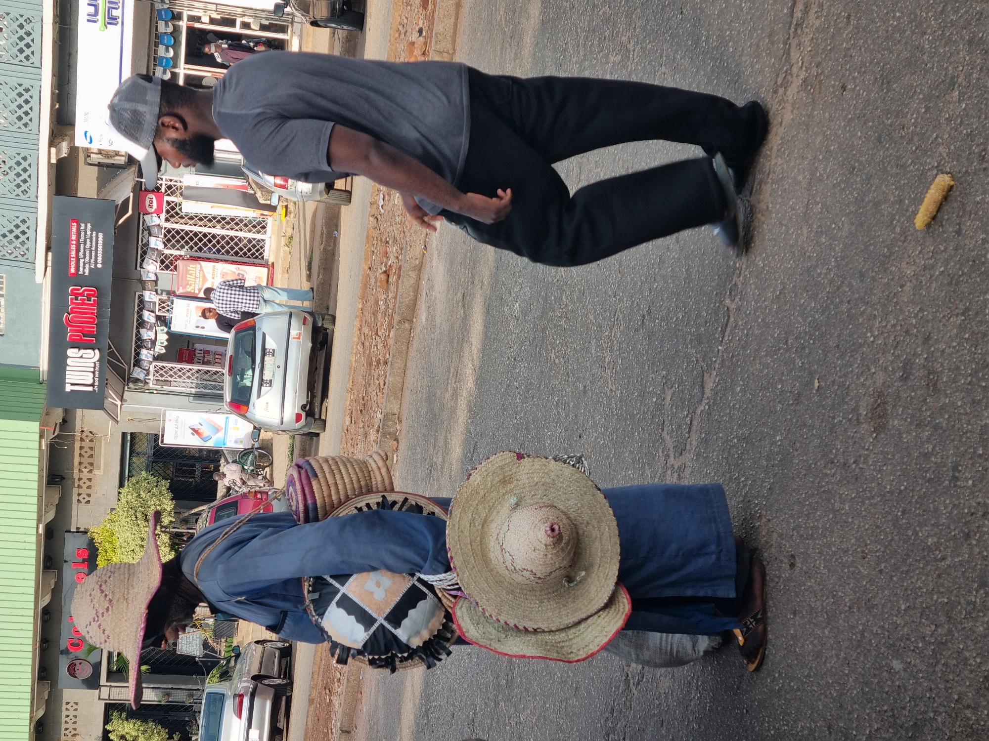 A vendor with traditional hats and baskets stands on a city street in Jos, Nigeria. The vendor is wearing a long blue garment and brown shoes. Various straw hats and baskets are stacked on their back. A corn cob lies on the street. A man wearing a grey t-shirt and black pants is walking past, looking at the goods. In the background, a car is parked outside of a store called Twins Phones, which sells mobile phones and accessories. Other cars and pedestrians are visible, suggesting a busy street scene.
