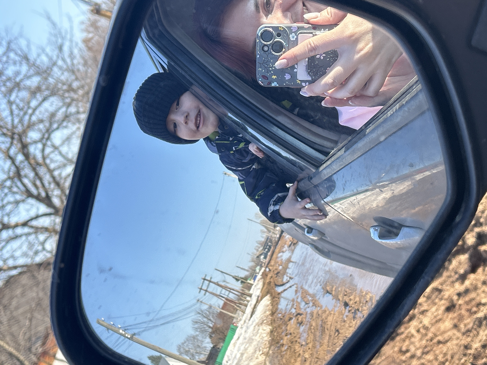 A view of a car's side mirror reflects a scene with two people. A woman is taking a photo with her phone, visible in the mirror's upper corner, and a child in a black beanie is looking out the car window. The reflection also shows a landscape with utility poles and patches of snow and dry vegetation. The setting is likely near the city of Фоки, Russia.