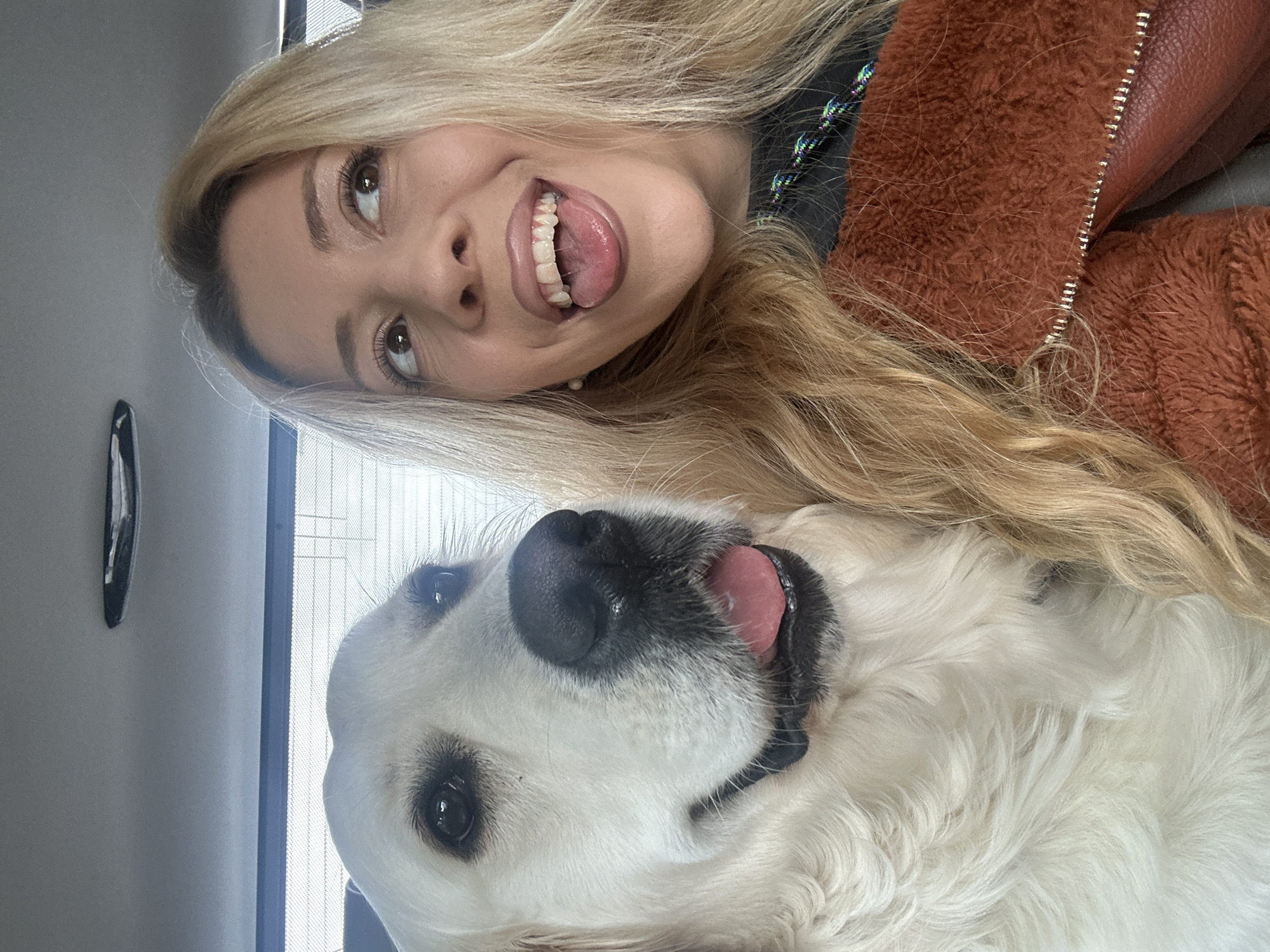 A selfie shows a young woman with long, blonde hair, and a white dog, possibly a Golden Retriever, indoors. The woman is in the upper half of the frame, with her face angled towards the camera, sticking her tongue out. The dog is positioned below her. The setting is most likely inside a vehicle, given the presence of a side window with visible blinds. There is no other evidence to suggest it is taken in the city of London, United Kingdom.