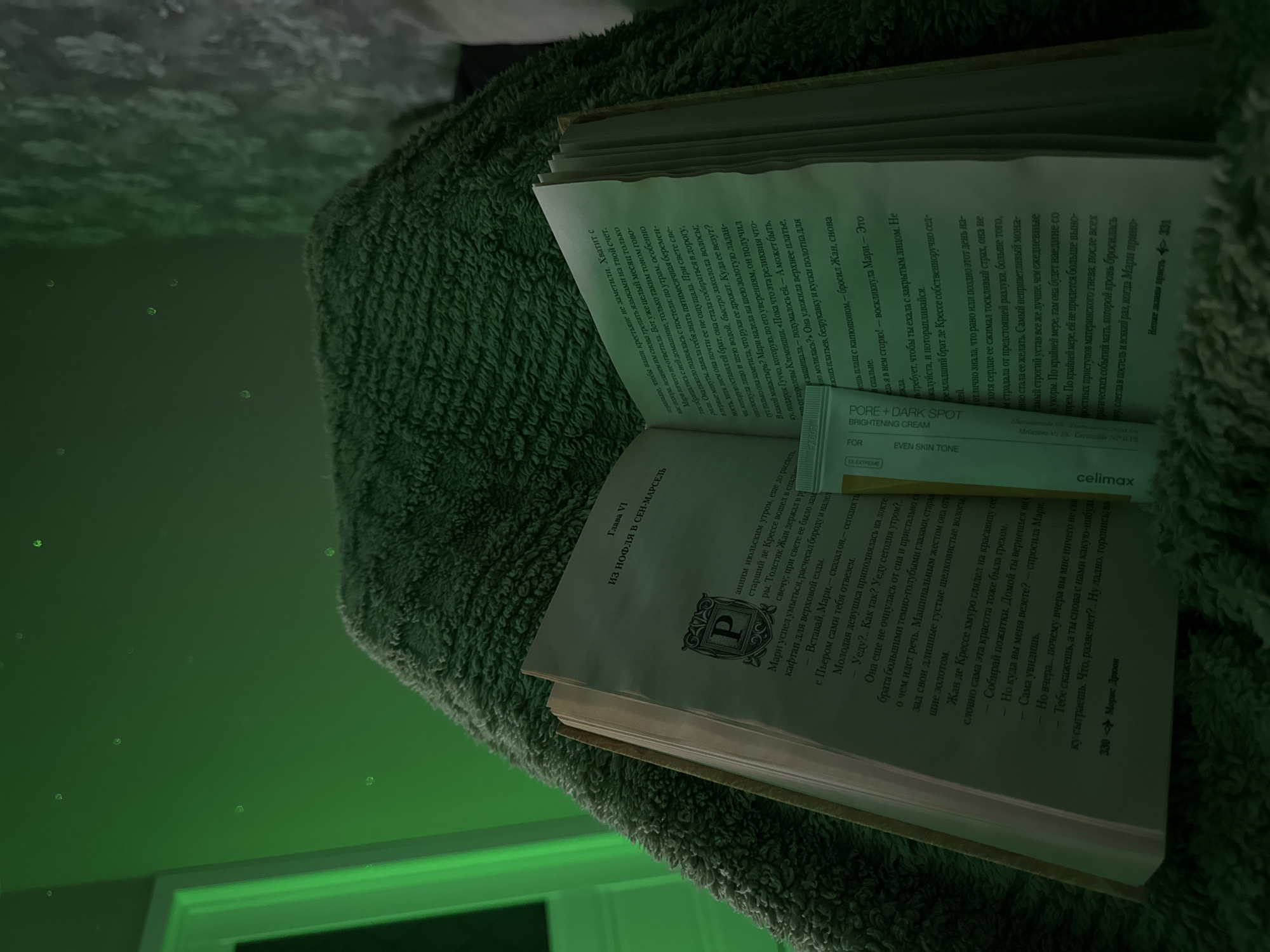Here is a detailed description of the image:
The image captures an open book resting on a soft, green, textured surface that appears to be a blanket or cushion. The book is written in Russian and has the title "pore + dark spot brightening cream" along with the brand name "celimax" visible on the page. The pages are filled with Russian text, and a black and white picture is noticeable on the left side of the book.
The book seems to be situated near a light source, evidenced by the greenish hue and highlights, particularly around the edges of the pages and the fluffy surface beneath. In the background, slightly out of focus, there's a hint of a wall or a structure with what appears to be decorative lighting or a light fixture, casting a subtle greenish glow. The angle and framing suggest a personal, intimate setting where someone might be reading and engaging with skincare or beauty products.