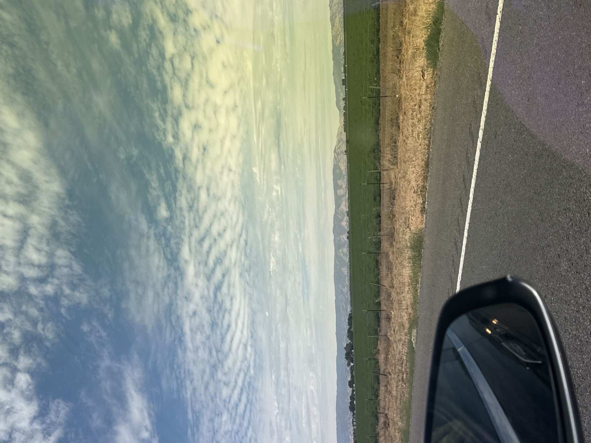 The image depicts a view from inside a vehicle, looking out towards a rural landscape. In the foreground, a portion of a black car's side mirror is visible on the left side of the frame, reflecting the surrounding environment. The road is asphalt, with a white dashed line indicating a lane divider. Beyond the road, a dry, grassy embankment transitions to a vibrant green field. A wire fence with metal posts runs along the edge of the field. In the mid-ground, a range of low, rolling hills can be seen, some with visible vegetation and others appearing more barren. The sky is filled with scattered white clouds against a blue backdrop, with a subtle color gradient from blue to yellow/green, possibly due to lighting or camera effects. The overall scene suggests an open, agricultural area with a wide sky, characteristic of the countryside. The location is identified as Dixon, United States of America.