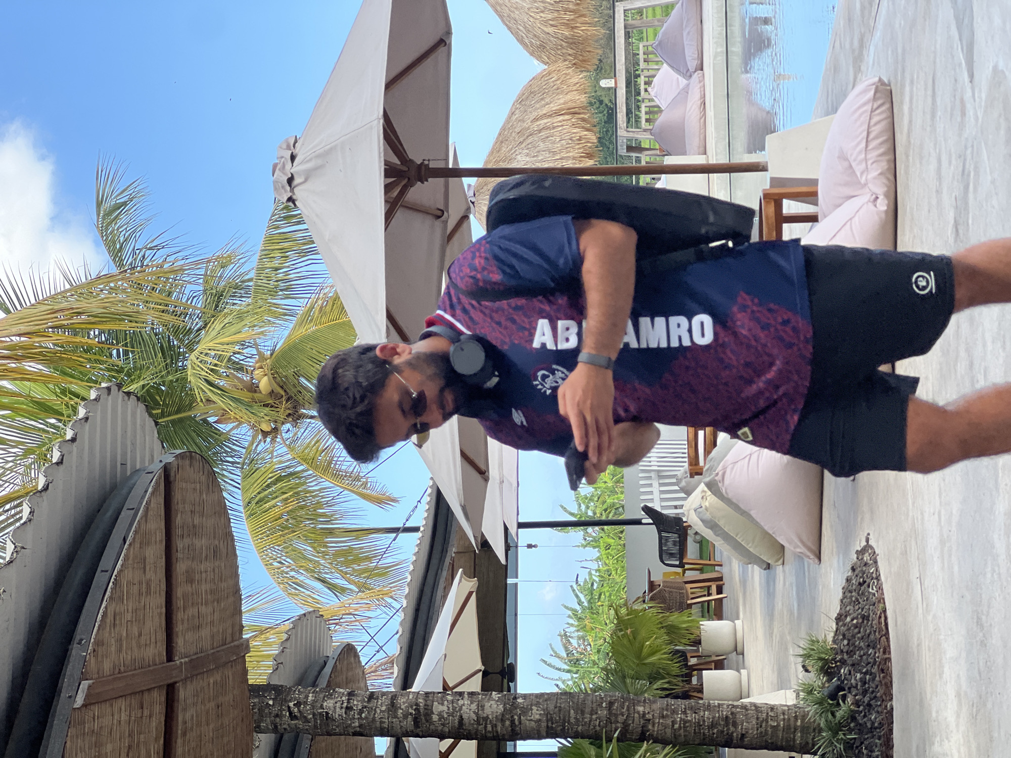 In North Kuta, Indonesia, a man with dark hair and a beard, wearing sunglasses, a dark blue and red jersey with white lettering that reads "ABRAMRO", black shorts, and a black backpack, is standing and looking at his mobile phone. He has headphones around his neck. The man is on a paved outdoor area near a swimming pool. In the background, there are palm trees, thatched umbrellas, white umbrellas, beanbag chairs, and some seating. The sky is blue with scattered clouds.