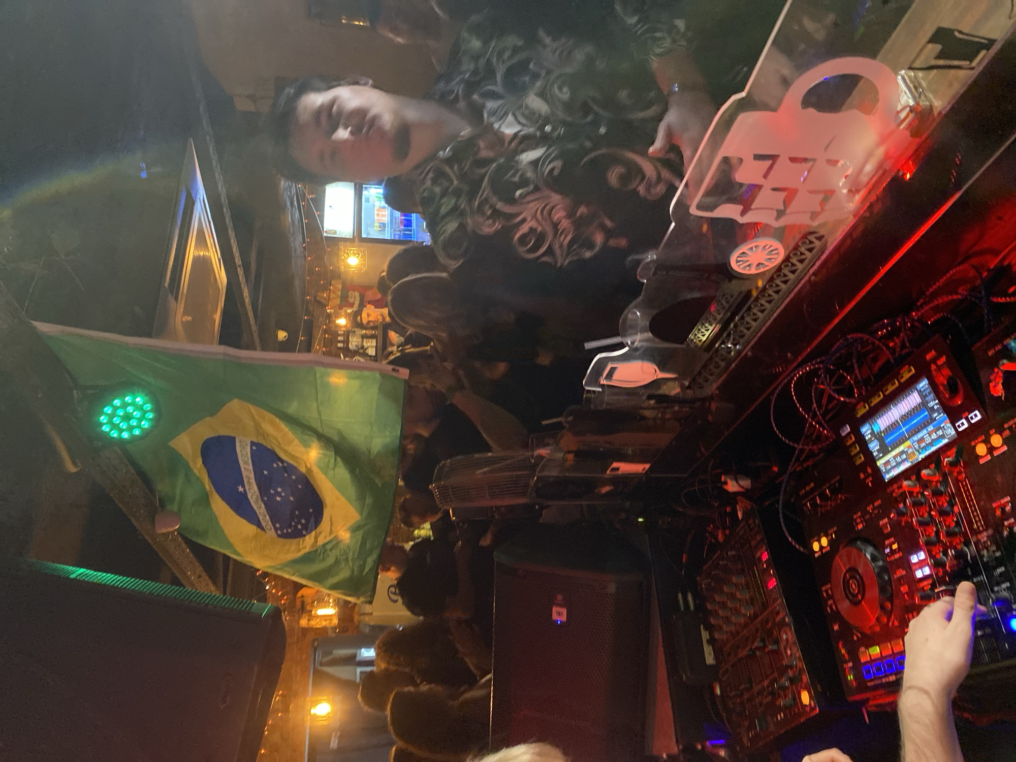 The image shows the interior of a nightclub or bar, possibly in Izhevsk, Russia. A DJ is performing, with a DJ mixing board visible, illuminated with red and blue lights. A person with dark hair and a patterned shirt is nearby, possibly a DJ assistant or club patron. A Brazilian flag is hanging in the background. A crowd of people is partially visible, suggesting a lively atmosphere. A speaker and a glowing neon sign with the number "777" are also visible. The location appears to be a dimly lit nightlife establishment.