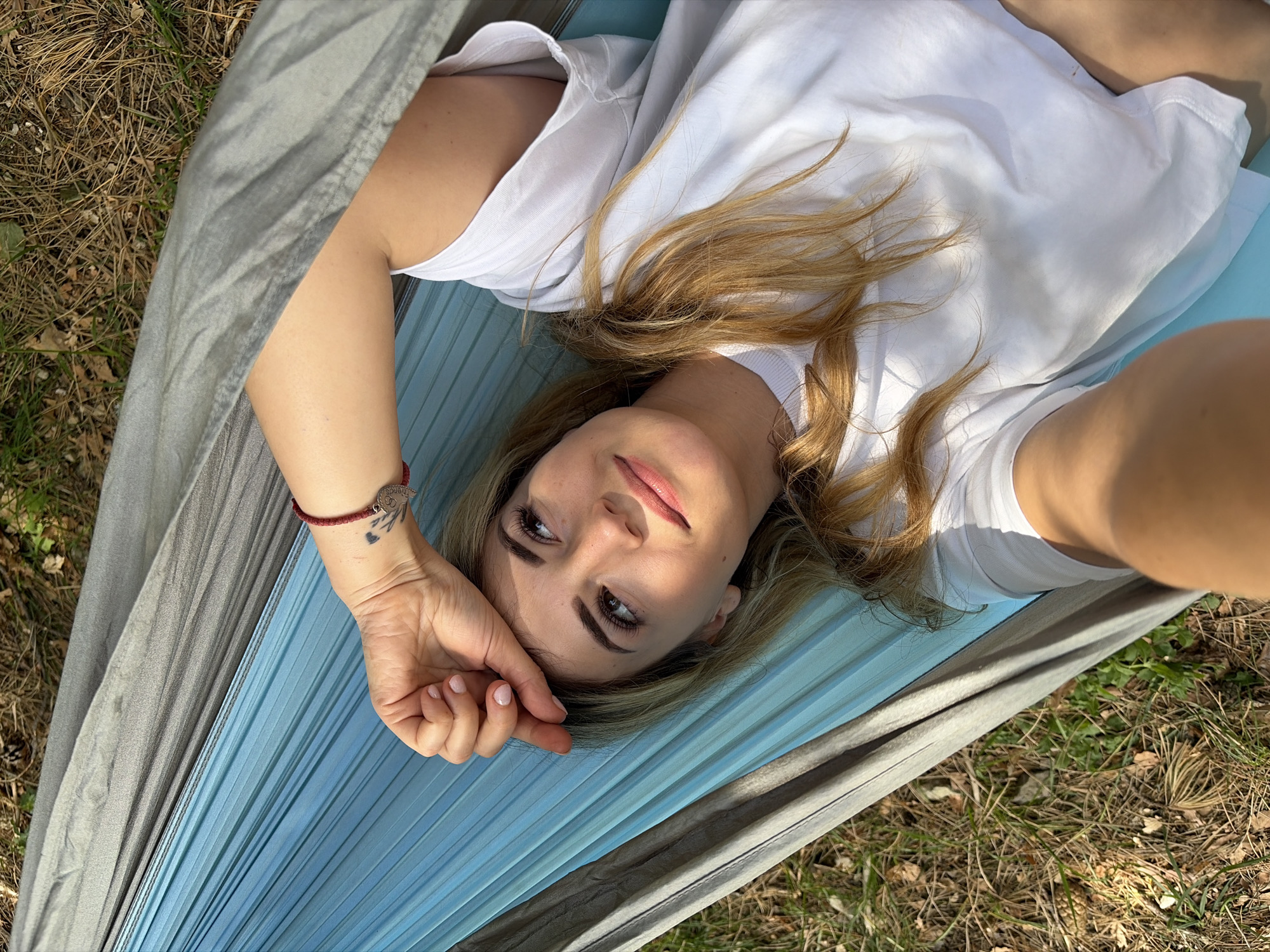The image depicts a fair-skinned female adult in a hammock, situated outdoors in Mali Bostur, Montenegro. She is lying on her back, wearing a plain white short-sleeved t-shirt. Her long, blonde hair is spread around her head. Her head is tilted slightly upwards, with her eyes open and directed towards the top-right of the frame. A subtle, serene smile is present on her face, and strong natural sunlight casts shadows across her features. Her left arm is bent, with her hand resting on her forehead, partially shielding her eyes from direct sunlight. A red woven bracelet adorns her left wrist, alongside a small tattoo featuring a heart-like shape with foliar elements. Her fingernails are painted a light pink. She is positioned within a two-toned fabric hammock, predominantly light blue in the central area and transitioning to a darker grey along its edges. The hammock fabric exhibits noticeable horizontal pleats. The ground visible beneath and around the hammock is covered with dry brown leaves, pine needles, and sparse patches of green vegetation, indicating a natural, unpaved outdoor setting. The photograph is taken from a high-angle perspective, looking down into the hammock.