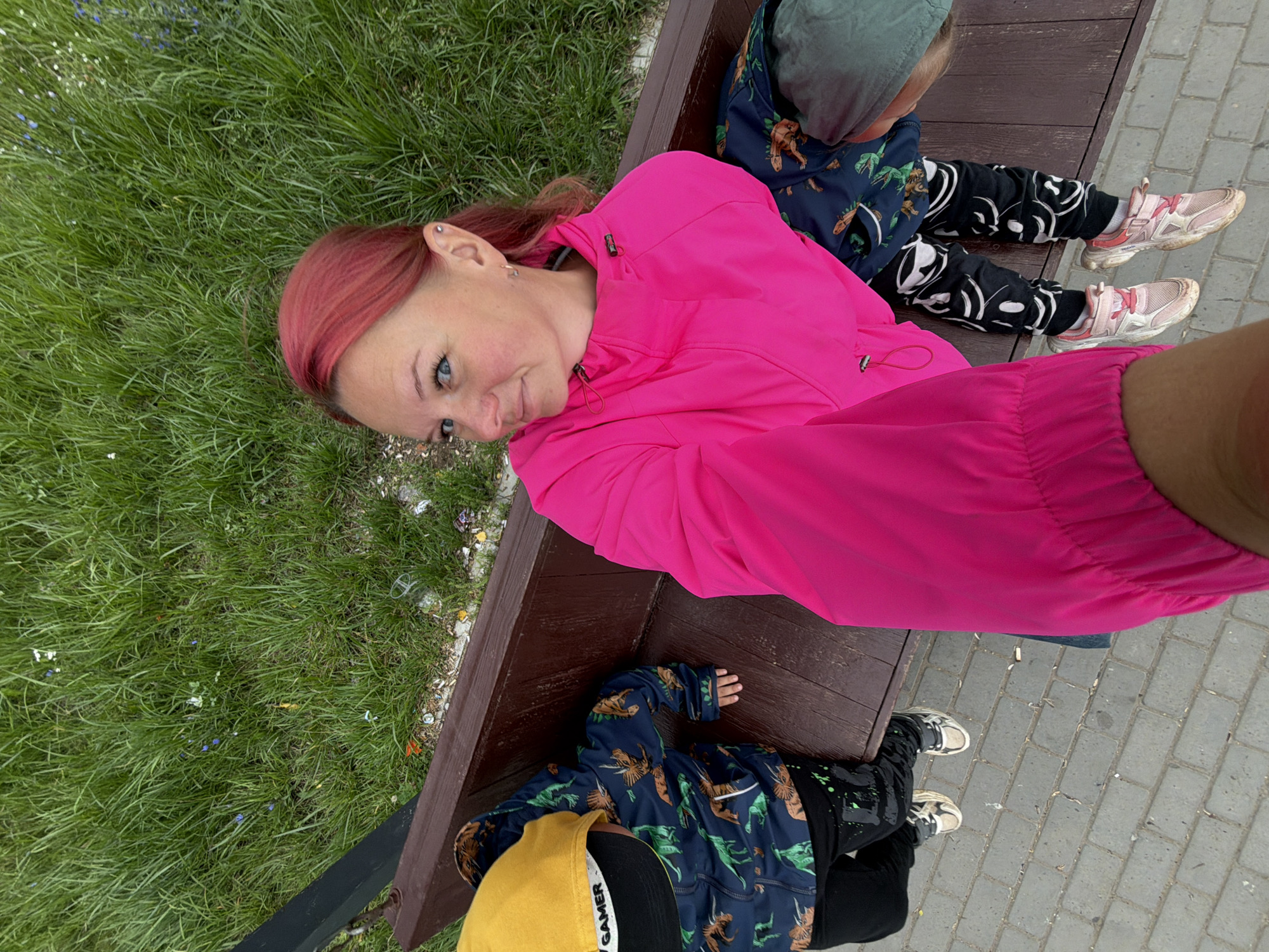 In Chaikovsky, Russia, a woman with bright pink hair is taking a selfie while sitting on a wooden park bench next to two young children. The woman is wearing a bright pink jacket and is looking at the camera with a slight smile. The child on the left is wearing a yellow hat with the word "GAMER" and a dark blue jacket with a dinosaur print. The child on the right is wearing a dark jacket with a dinosaur print and black pants with white smiley faces. The bench is situated on a paved walkway with green grass and small wildflowers in the background. The scene appears to be outdoors in a park or recreational area.