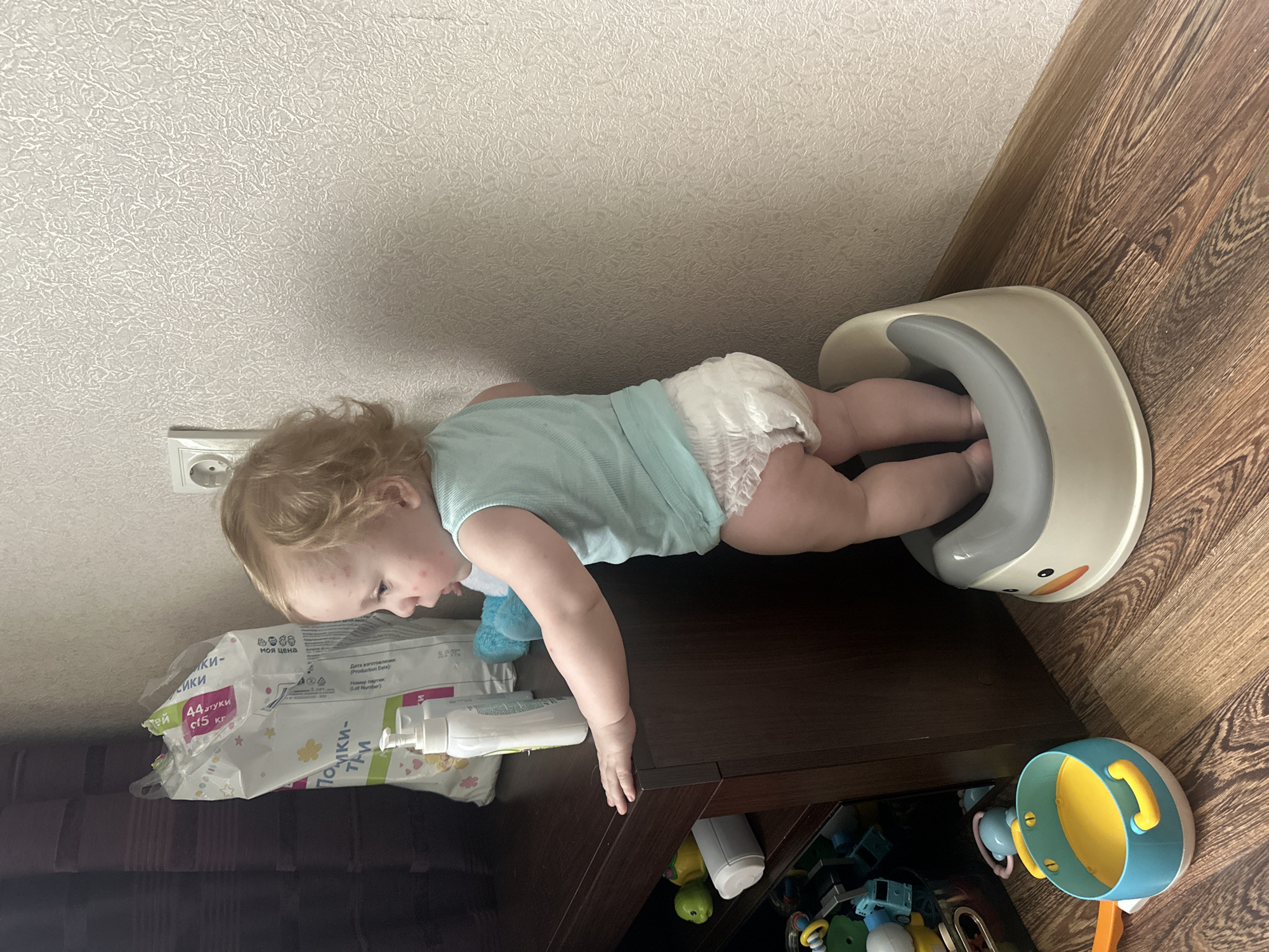 The image depicts a toddler wearing a diaper standing in a potty training seat. The potty seat is white with a gray rim and a duck design on the front. The child has blonde hair and appears to be looking to the left. The child is standing on a wooden floor in what appears to be a residential setting. To the left of the child, there is a dark brown dresser or cabinet. On top of the cabinet, there is a large white bag, possibly containing diapers, and a white pump bottle. On a shelf below the cabinet, there are several toys, including a blue and yellow toy pot. The background features a textured, light-colored wall with a white electrical outlet. The scene is lit by artificial light. The image was taken in a room in Sayanogorsk, Russia.