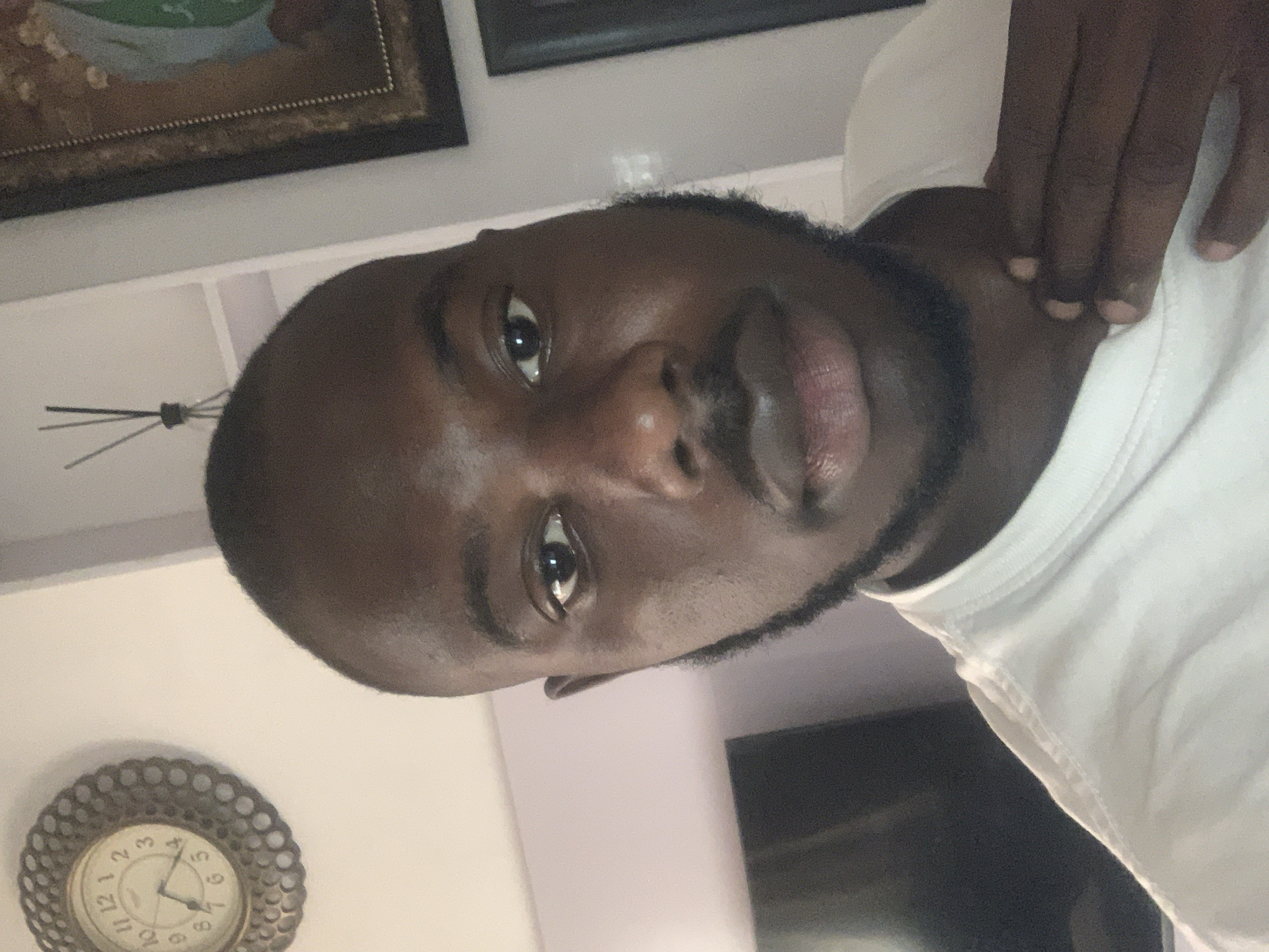 A selfie captures a dark-skinned man indoors. He is wearing a white shirt and has short, dark hair with a beard. His right hand is touching his neck. In the background, there are pictures on the wall, a shelf, a television, and a clock. The location is likely a residential area in Jos, Nigeria.