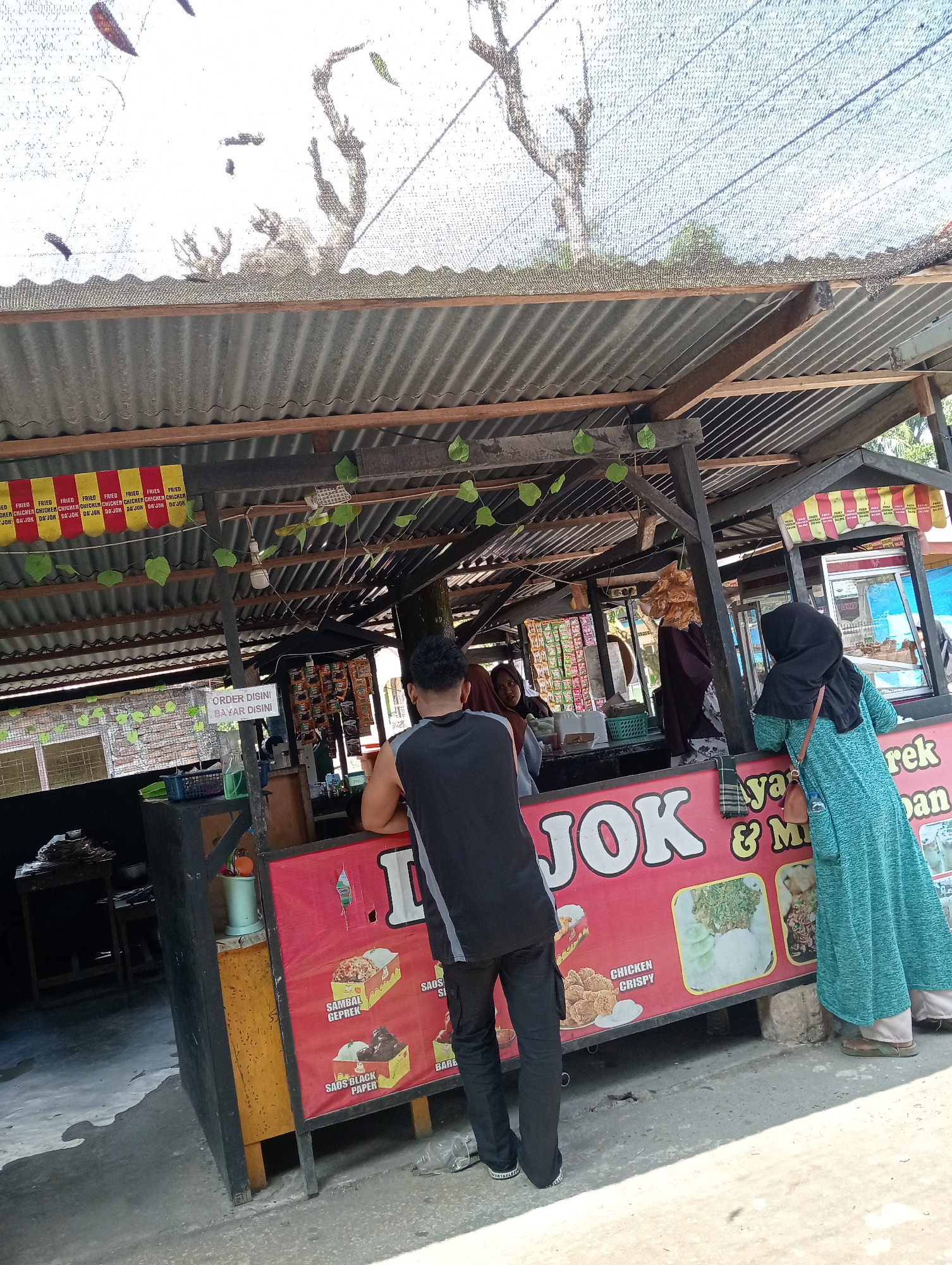 The image depicts an outdoor food stall in the Campago area of Indonesia. A corrugated metal roof shelters the stall, supported by wooden beams. Beneath the roof, a red banner displays the text "JOK" in large white letters, suggesting it's the name of the establishment or a product. The banner also features images of food, including fried chicken and rice dishes, with accompanying text like "CHICKEN CRISPY," "SAMBAL GEPREK," and "SAOS BLACK PAPER."
Several people are present. In the foreground, a man in a black sleeveless shirt and dark pants faces away from the camera, leaning on the counter. To his right, a woman wearing a blue-green hijab and dress stands, interacting with someone inside the stall. Another woman, wearing a dark headscarf, is visible behind the counter, seemingly serving customers. Various food products and items are displayed on shelves behind the counter, including packets of snacks and drinks. A sign that reads "ORDER DISINI BAYAR DISINI" (Order here, pay here) is also visible.
The ground appears to be paved with concrete. The overall environment suggests a casual, open-air dining or takeaway setting typical of local food vendors. The lighting indicates it is daytime with bright sunlight.